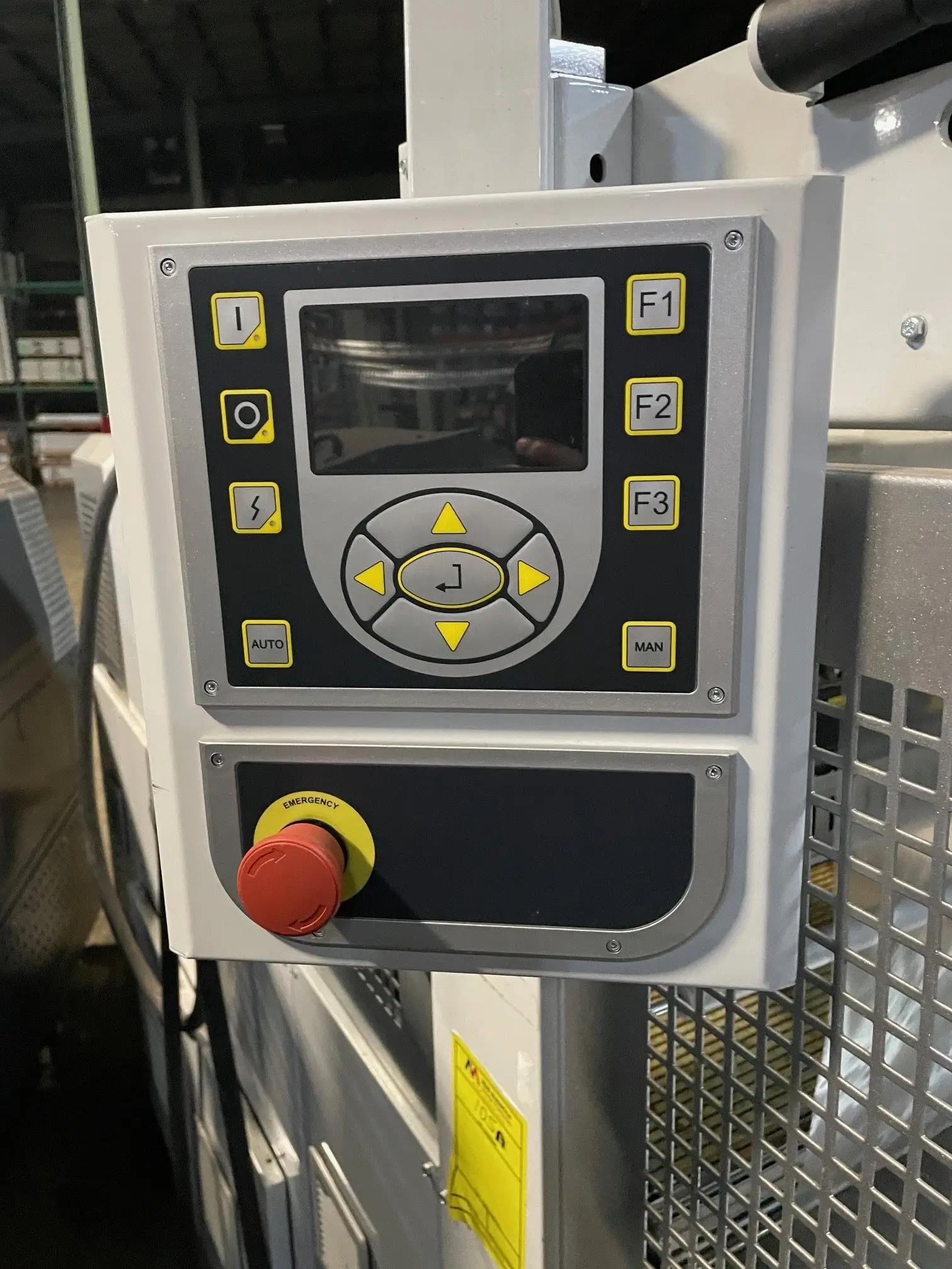 Close-up of an industrial control panel with a display screen, various buttons, and a red emergency stop button, located in a warehouse setting.