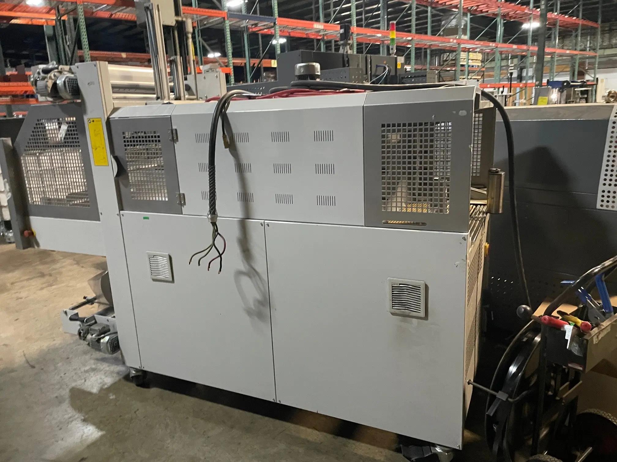 Industrial machinery in a warehouse setting, featuring a large grey metal machine with control panels and cables, surrounded by industrial shelving.