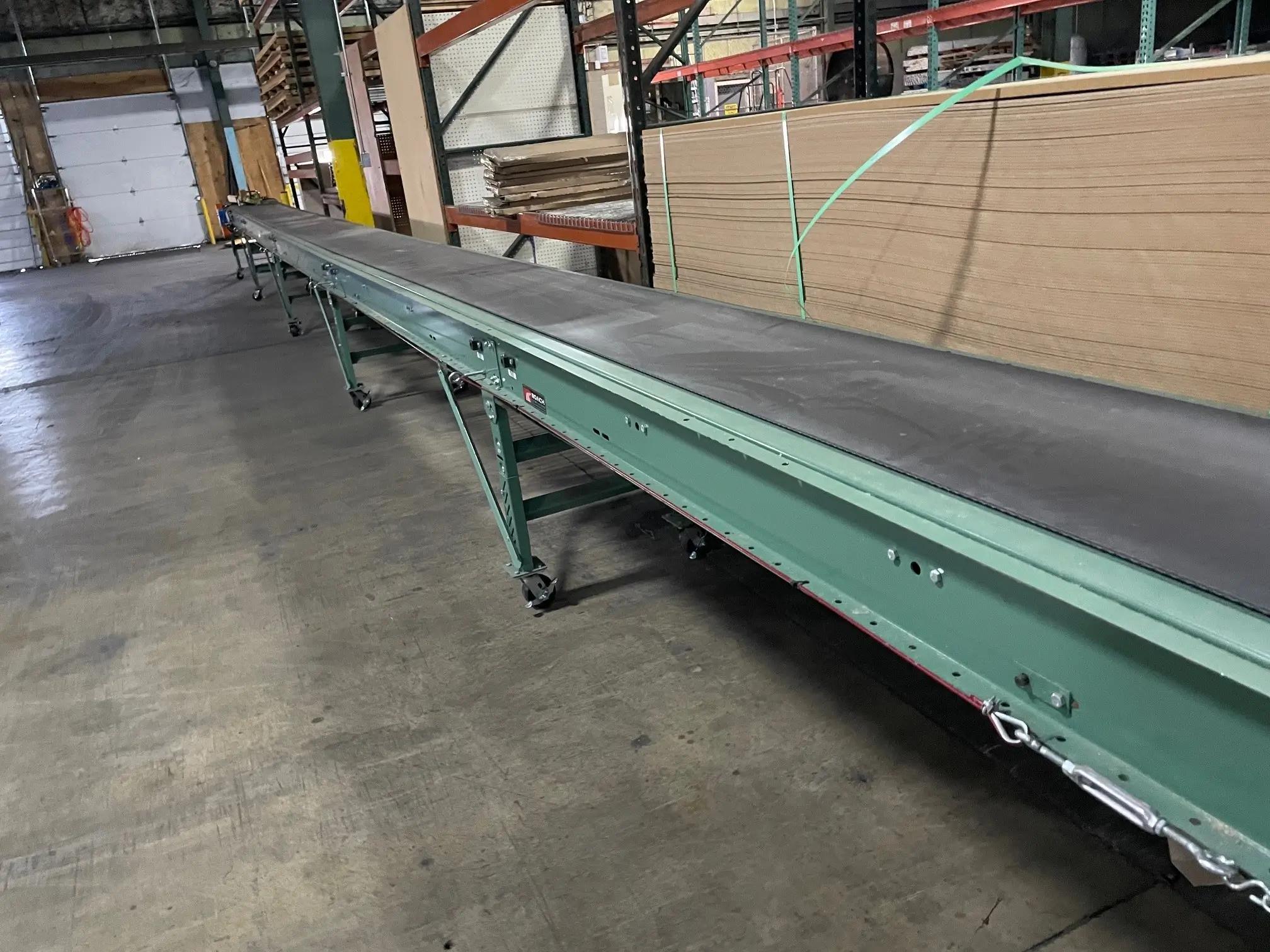 Conveyor belt system in a warehouse setting with stacked cardboard panels on shelving.