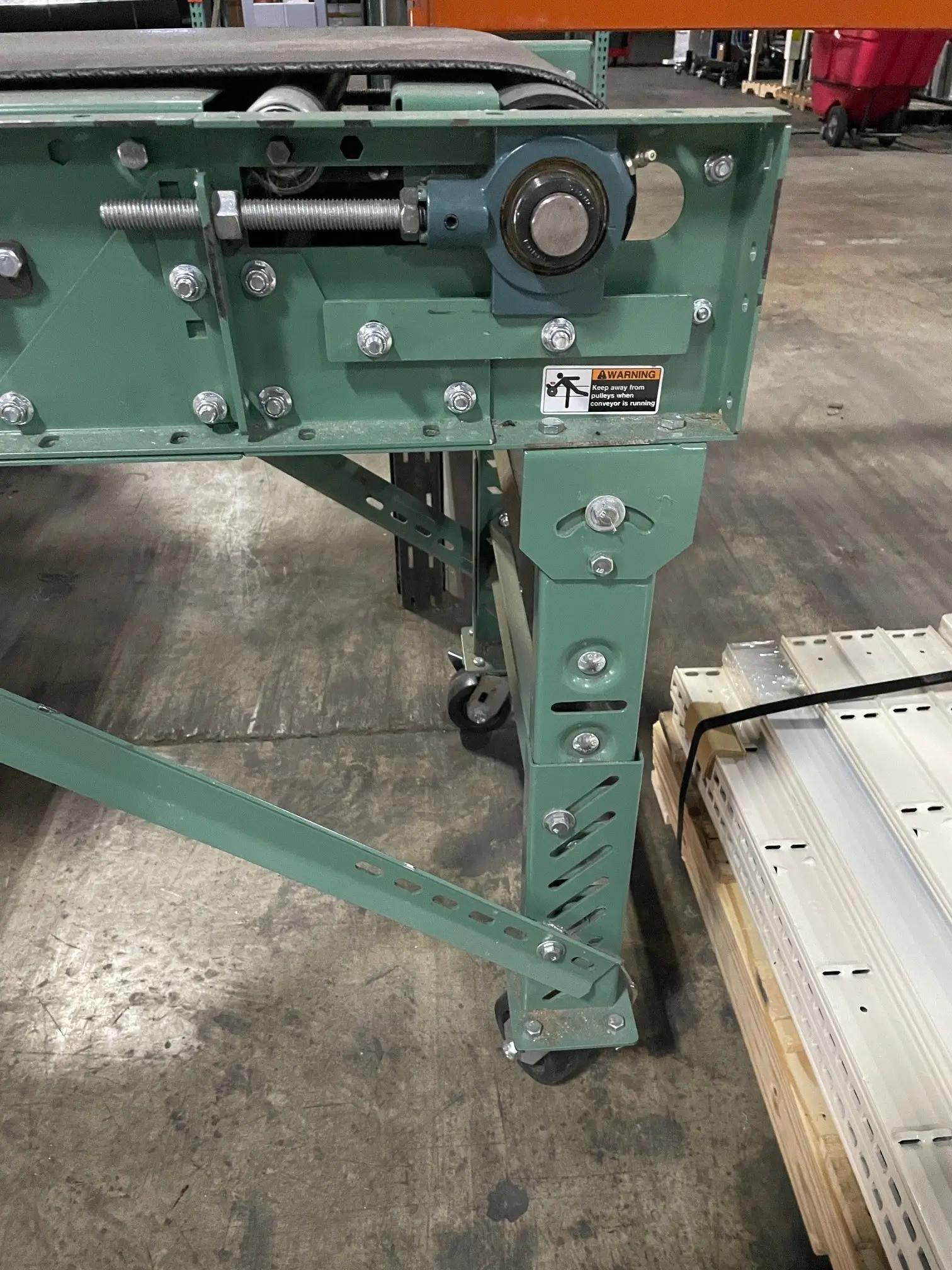 Close-up of a green metal conveyor system with rollers, bolts, and wheels in a warehouse setting. Part of a pallet is visible next to it.