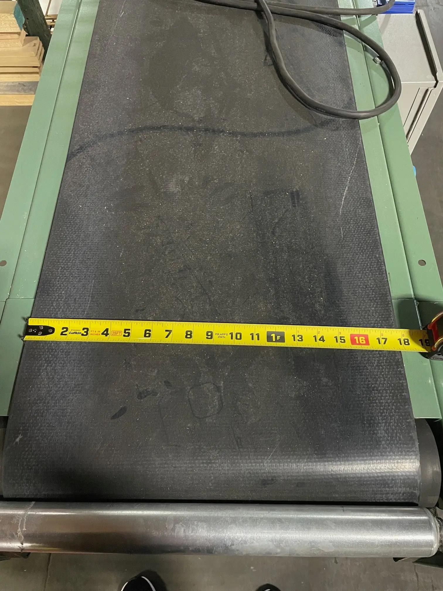 An industrial conveyor belt with a measuring tape stretched across its width, indicating a measurement of about 16 inches, in a workshop setting.