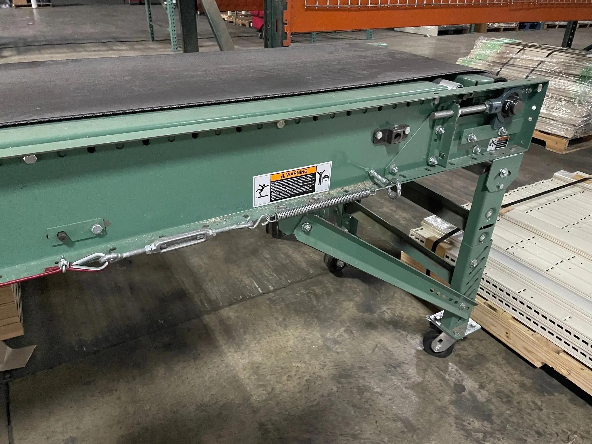 A green conveyor belt with safety warning labels in a warehouse setting, featuring rollers and a black belt.