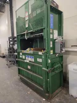 A green industrial compactor in a warehouse, used for compressing cardboard and other materials, with labels and controls on its front panel.