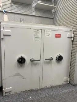 A large white double-door safe with combination locks and handles, located in a room with gray carpet and tiled walls. Shelves and a roller shutter are visible in the background.