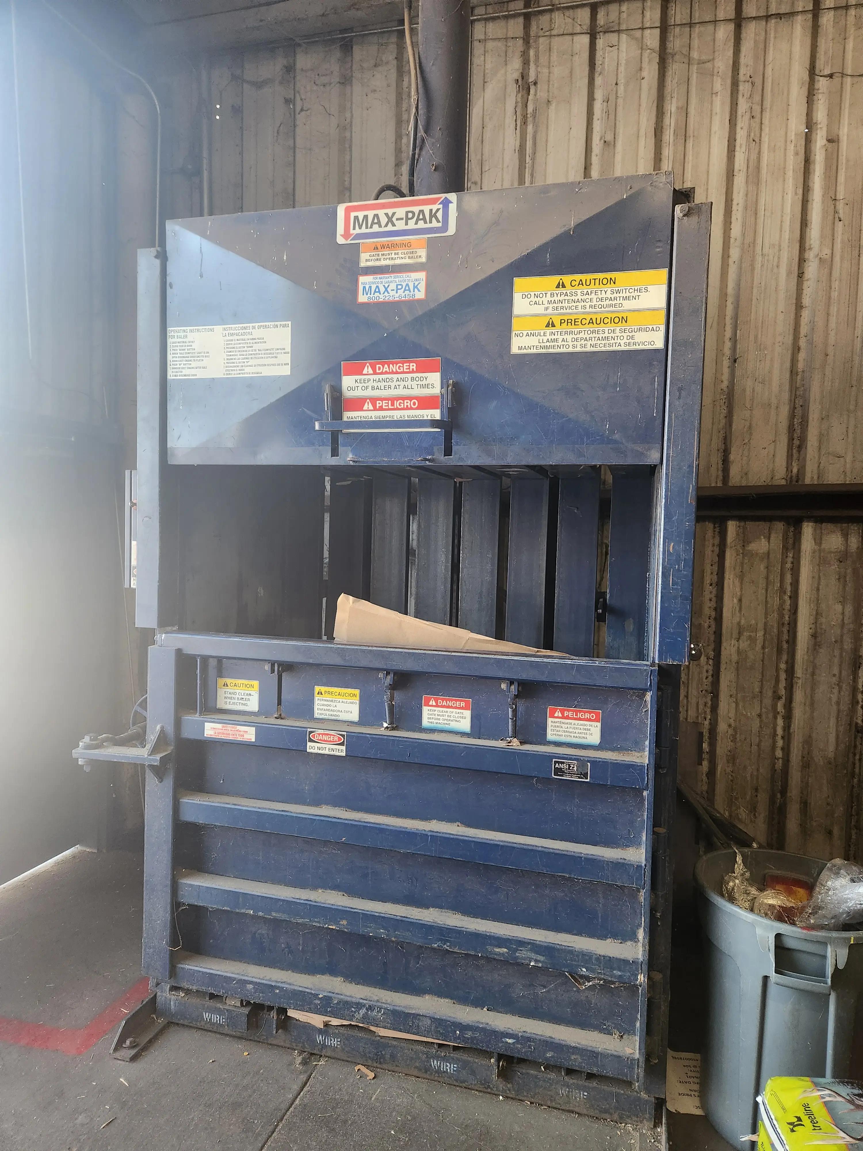 Image of a blue MAX-PAK baler in an industrial setting, featuring safety warning labels in English and Spanish. The baler displays instructions and caution signs emphasizing safe operation and the importance of keeping hands clear.