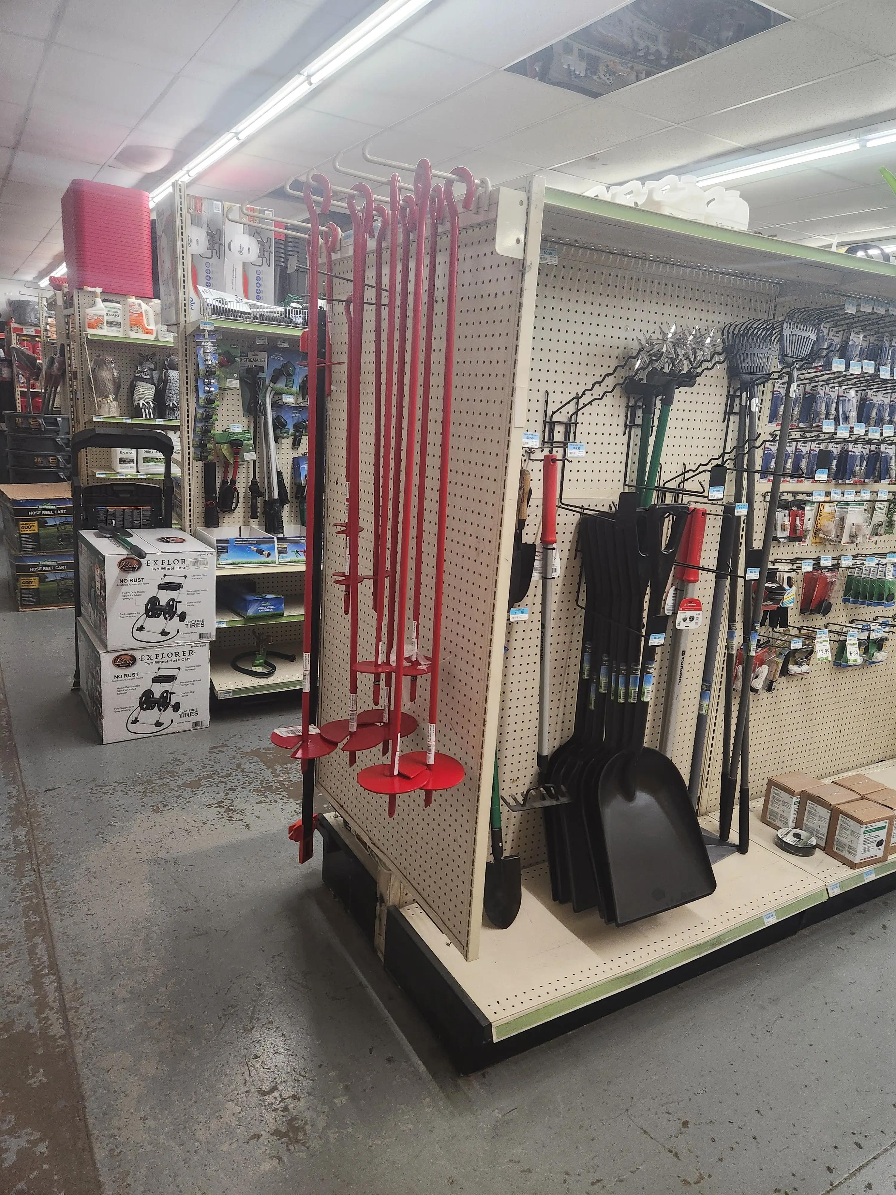 A hardware store aisle with various tools and equipment. Red augers are prominently displayed on a pegboard, alongside shovels and gardening tools. Shelves in the background hold boxes and additional supplies.