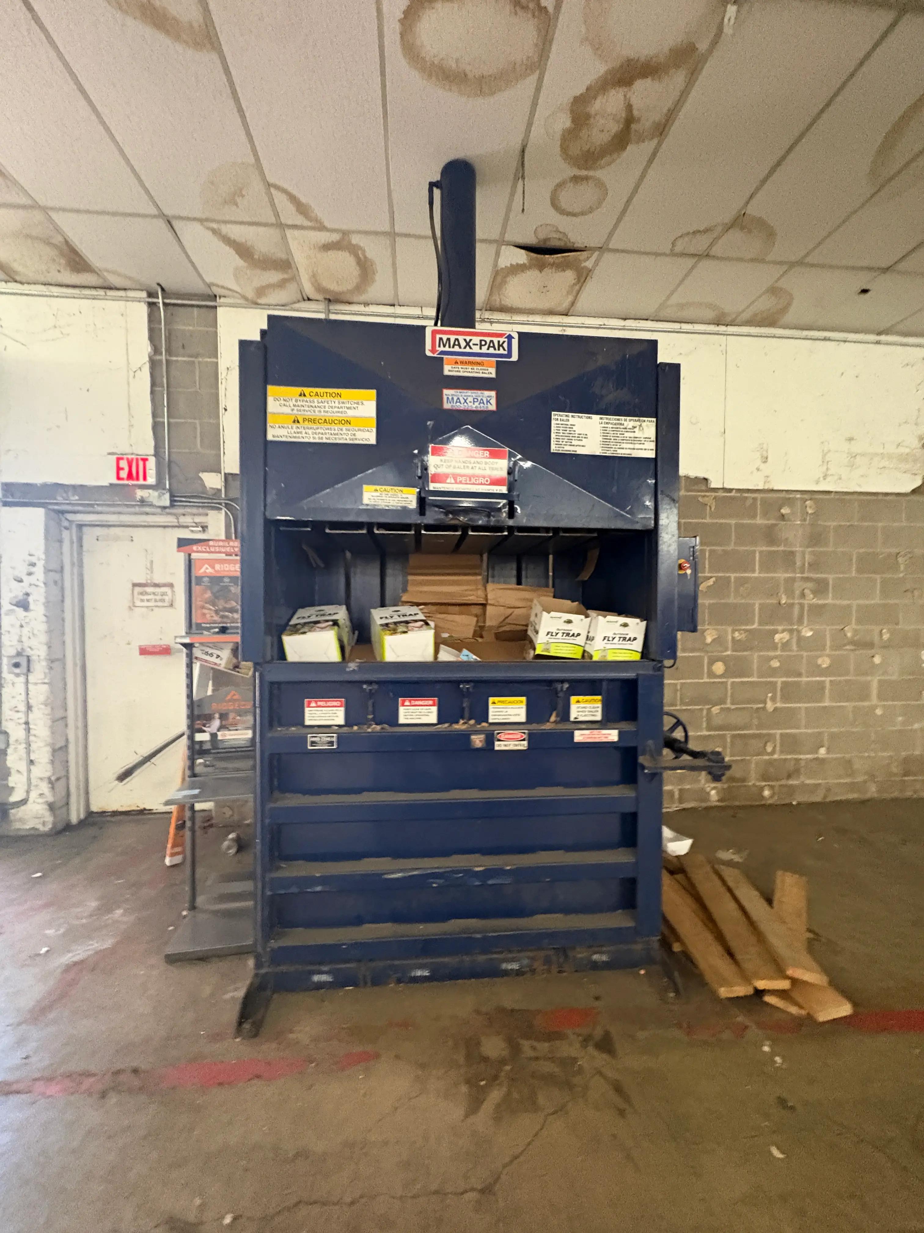 Industrial cardboard baler machine in a warehouse setting with safety signs and cardboard boxes