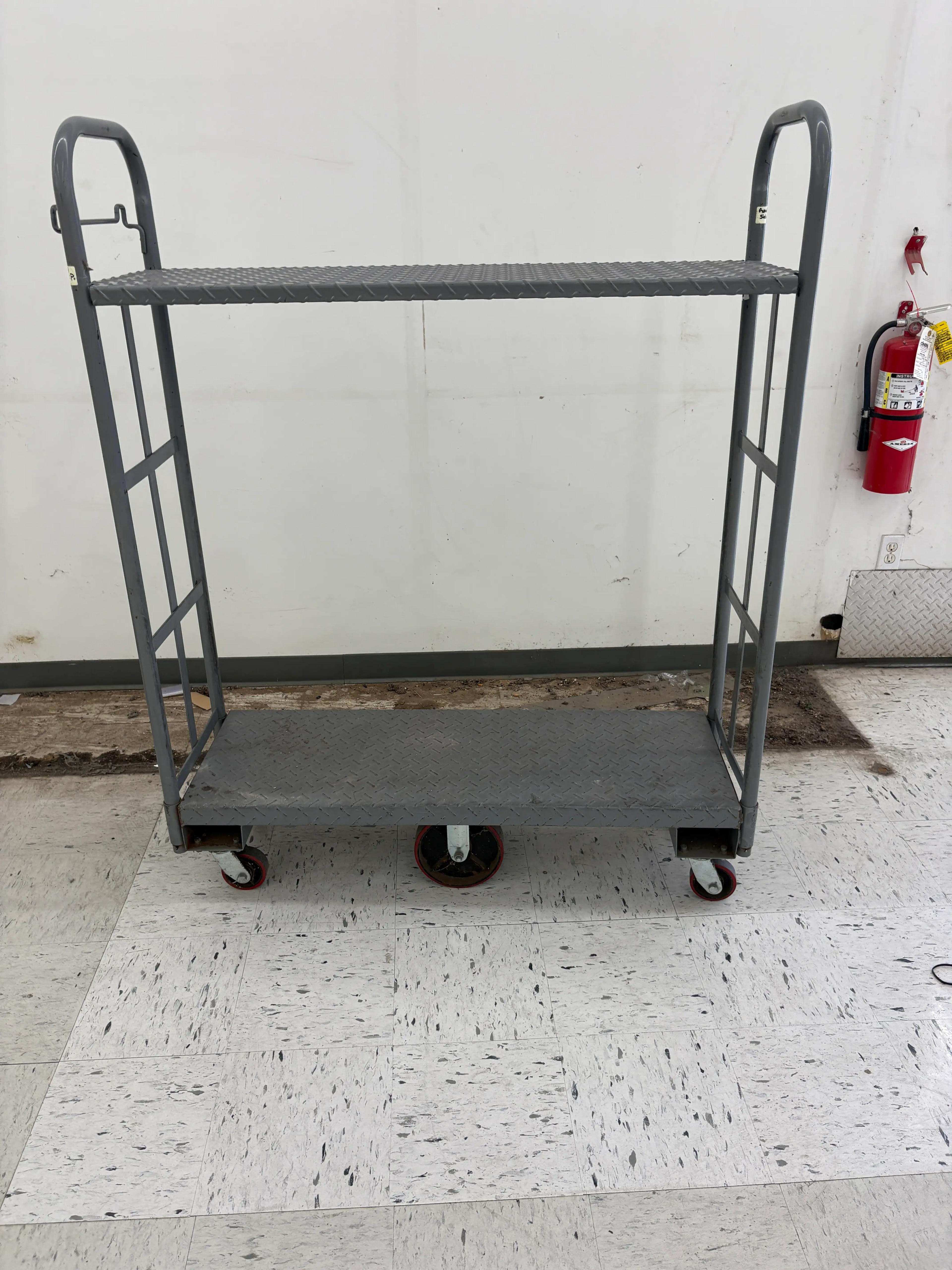 Gray metal cart with two shelves and red wheels, positioned in an industrial setting with a fire extinguisher in the background.