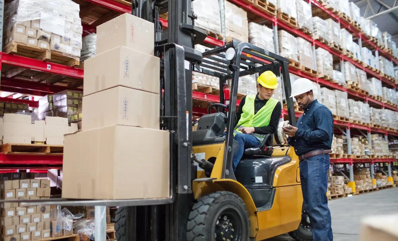Two workers in a warehouse with one operating a forklift and the other checking a document. Both are wearing hard hats and safety vests.