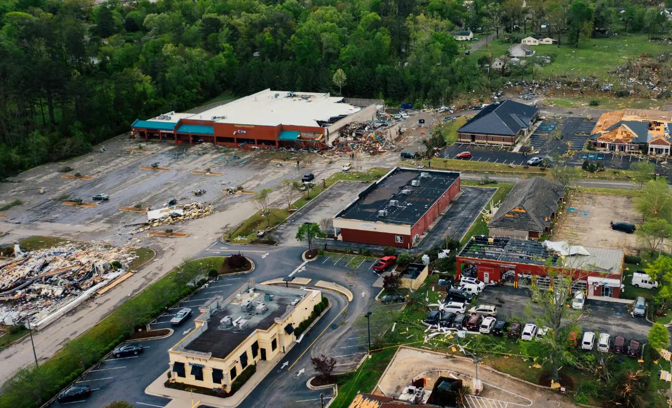 Aerial view of a neighborhood showing severe tornado damage, with debris scattered around parking lots and buildings partially destroyed.