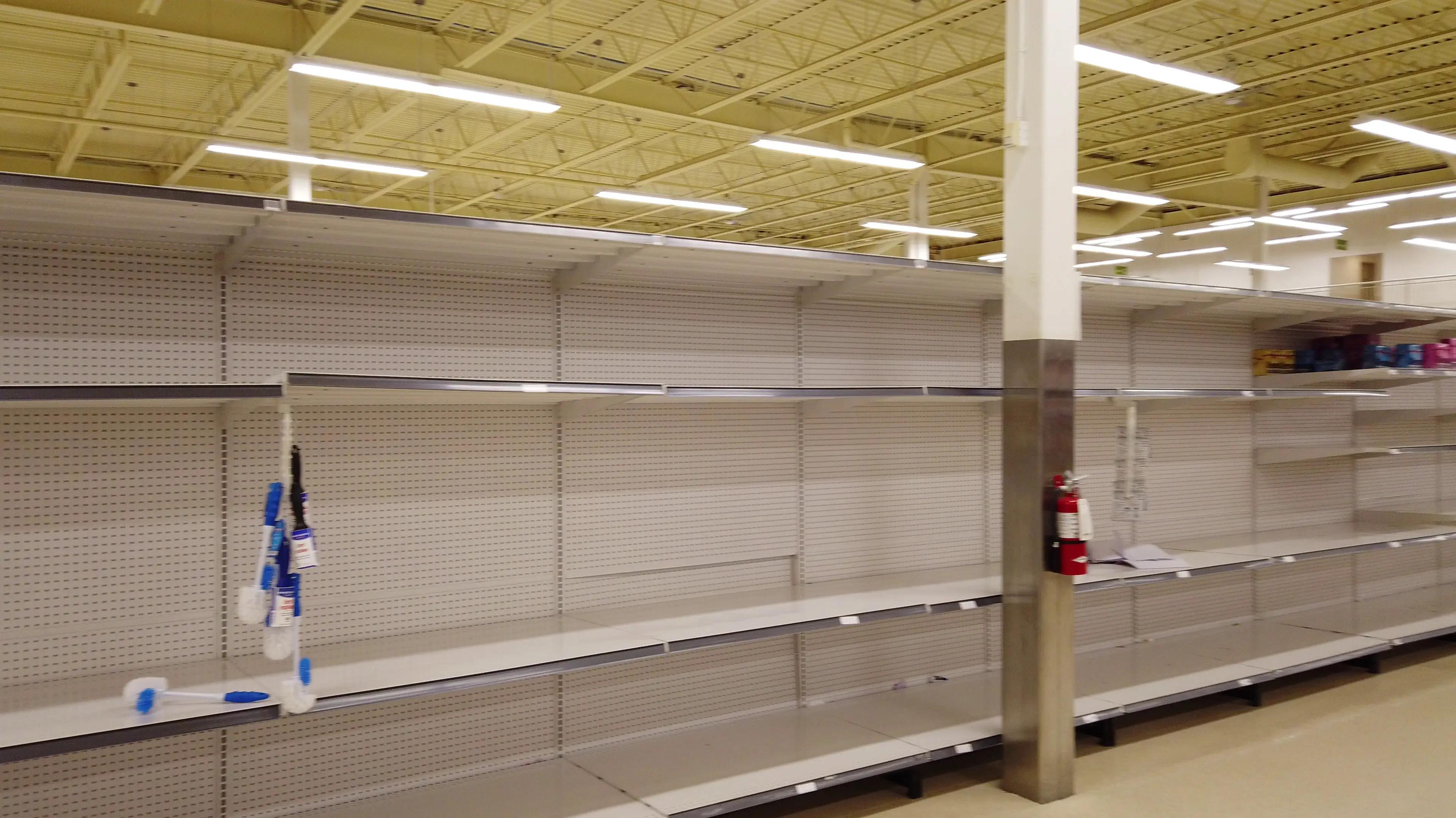 Image of empty shelves in a retail store, indicating a stock shortage, with few remaining cleaning brushes and a fire extinguisher visible.