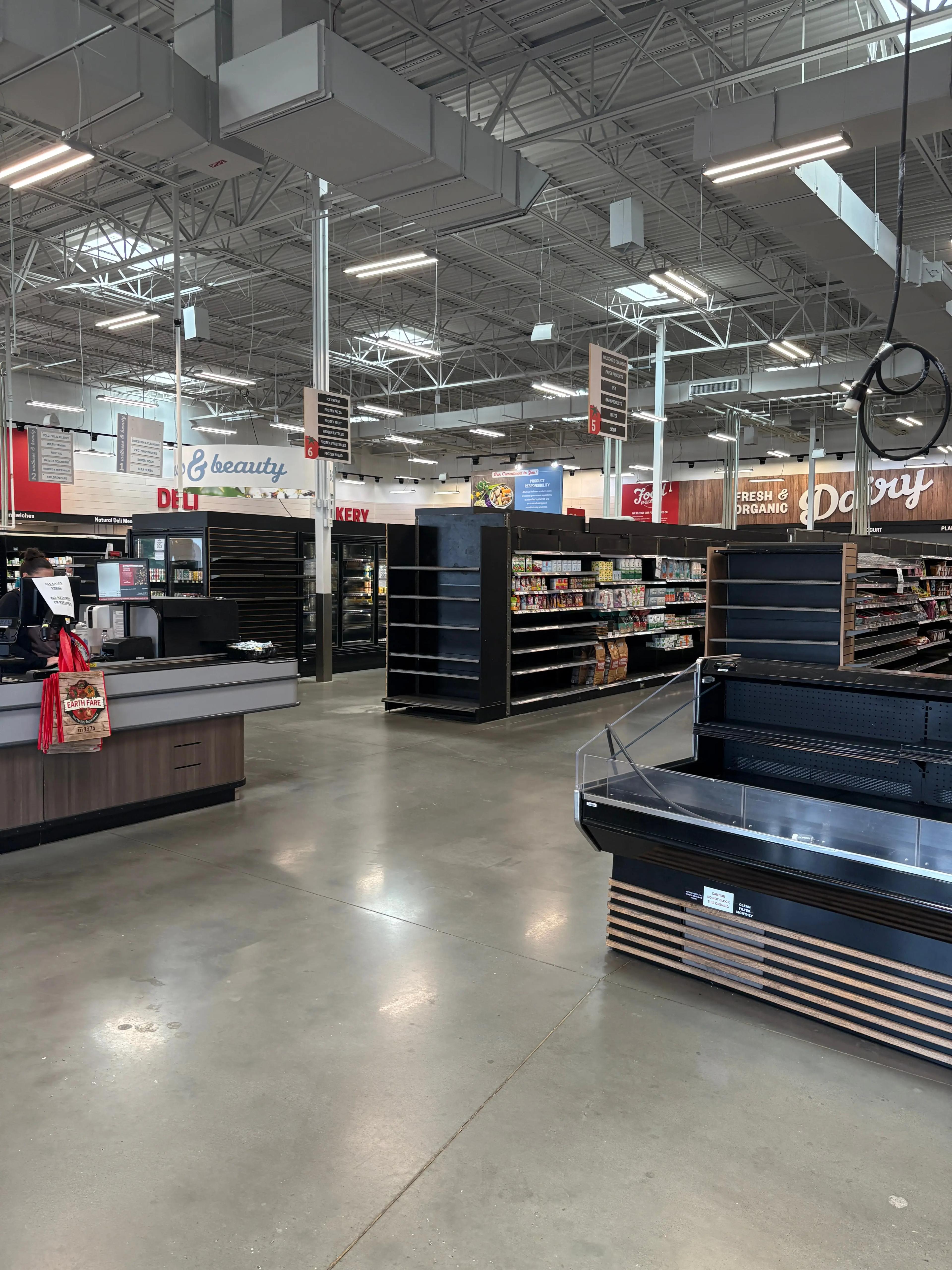 Interior view of a grocery store with empty shelves, modern lighting, and visible signage in sections like beauty, deli, and dairy.