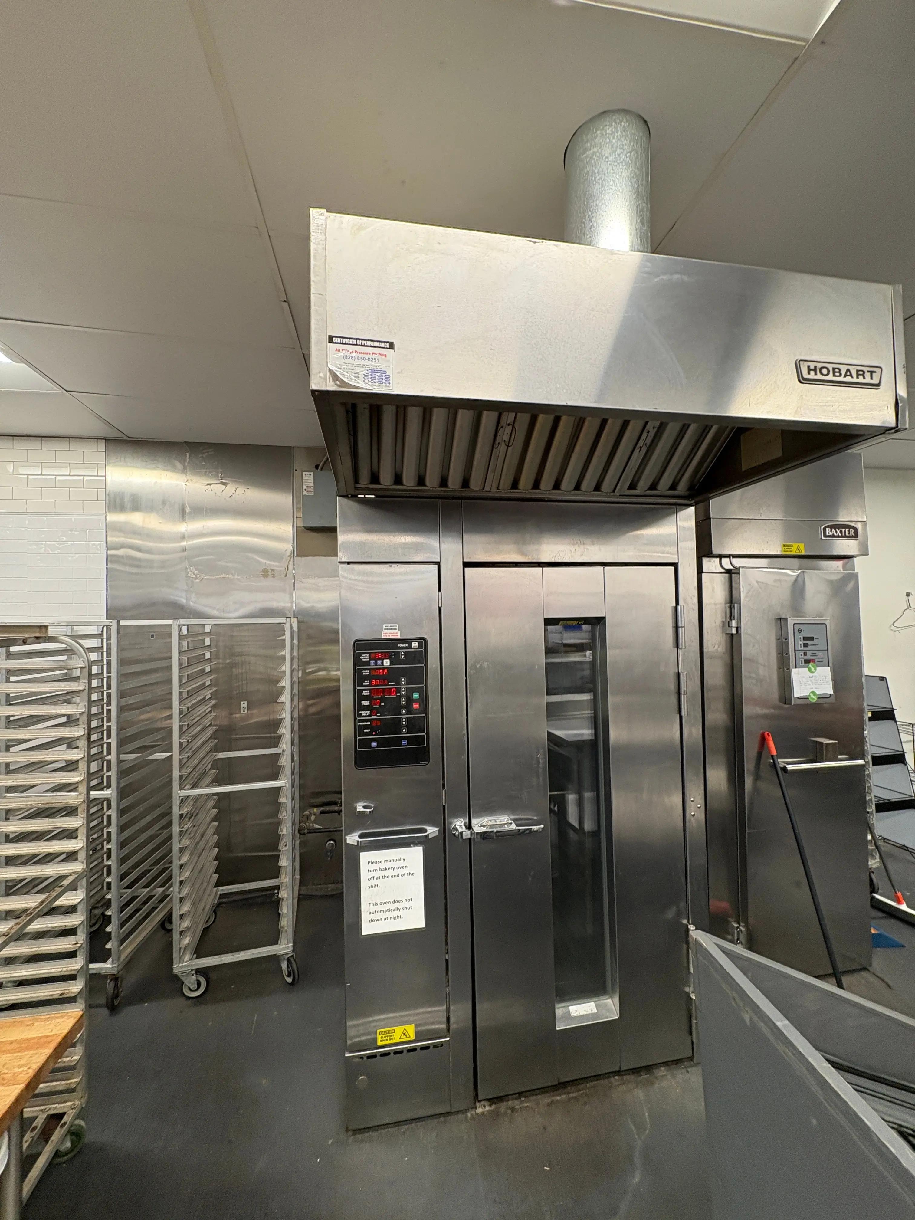 Commercial kitchen featuring a large stainless steel bakery oven labeled "Hobart" and "Baxter," with nearby metal racks and a caution sign advising manual shutdown of the oven at the end of shifts.