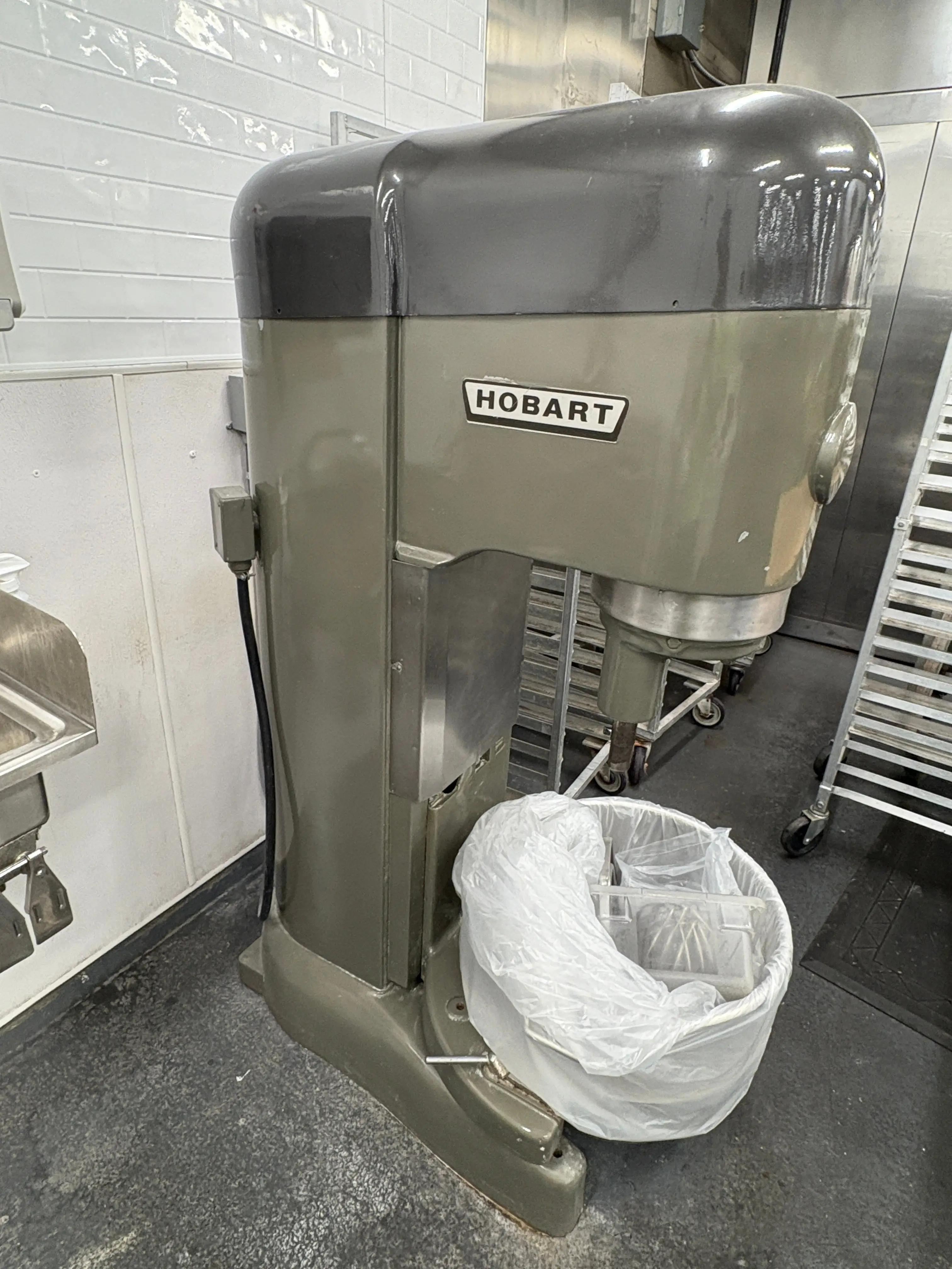 A large industrial Hobart mixer in a commercial kitchen, with a trash bin at its base. The mixer is gray and black, and the kitchen features white tiled walls and metal shelving in the background.