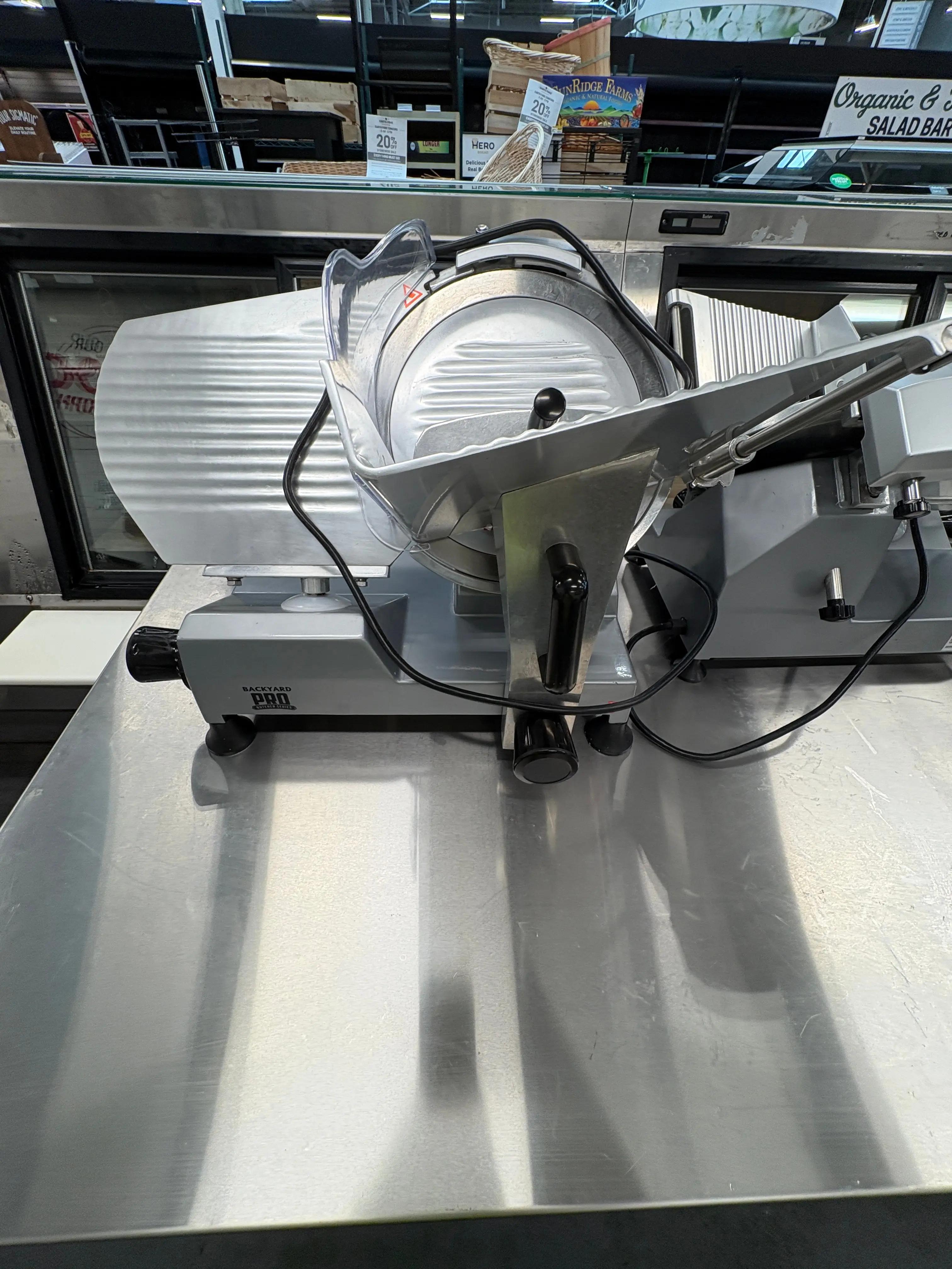 A commercial meat slicer branded "Backyard Pro" on a stainless steel counter in a store setting, with various products and signs visible in the background.