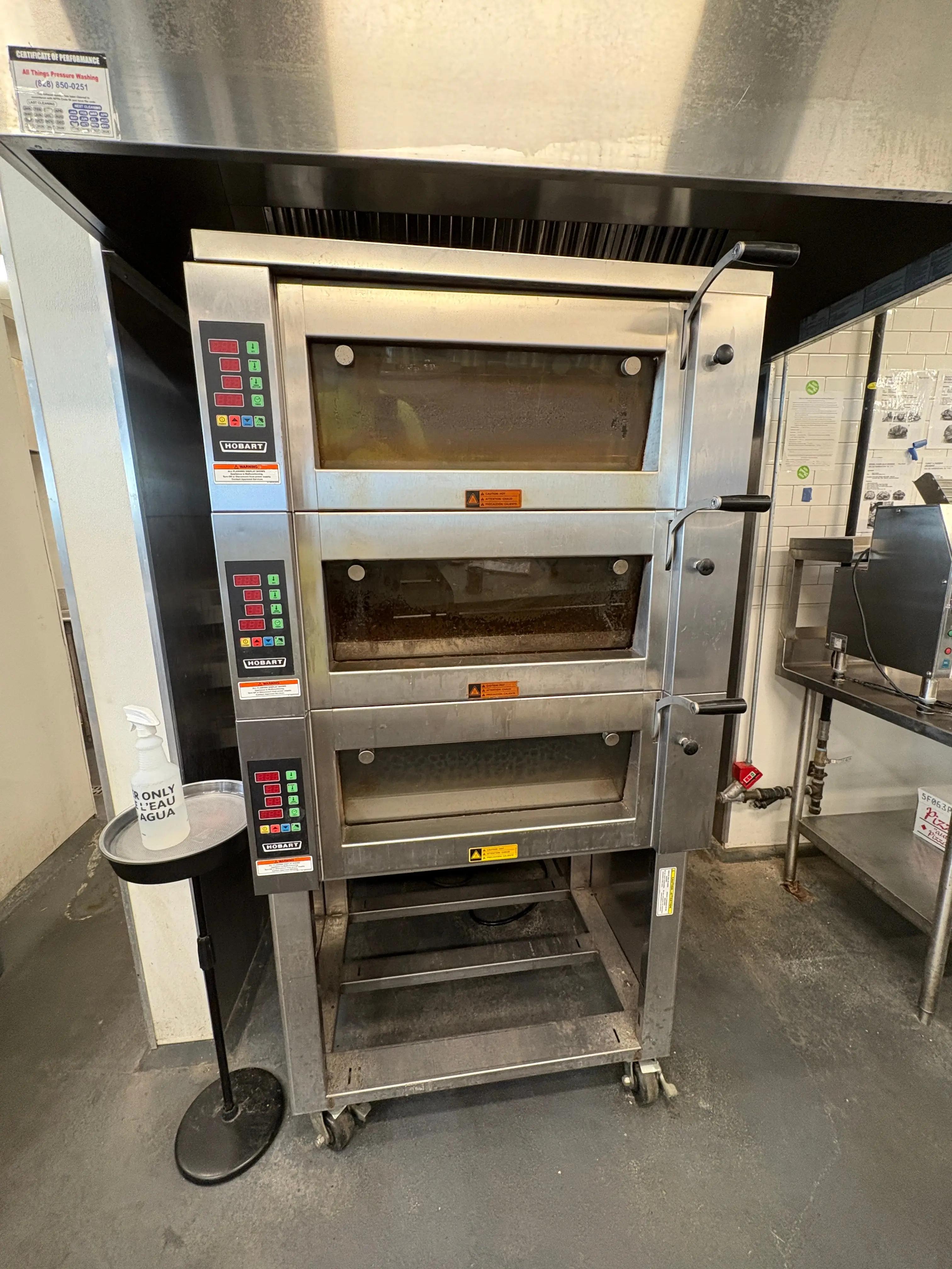 Industrial kitchen with a Hobart oven featuring three stacked compartments and control panels. A bottle labeled "ONLY CLEAN AGUA" is on a stand nearby. Stainless steel surfaces and equipment are visible in the background.