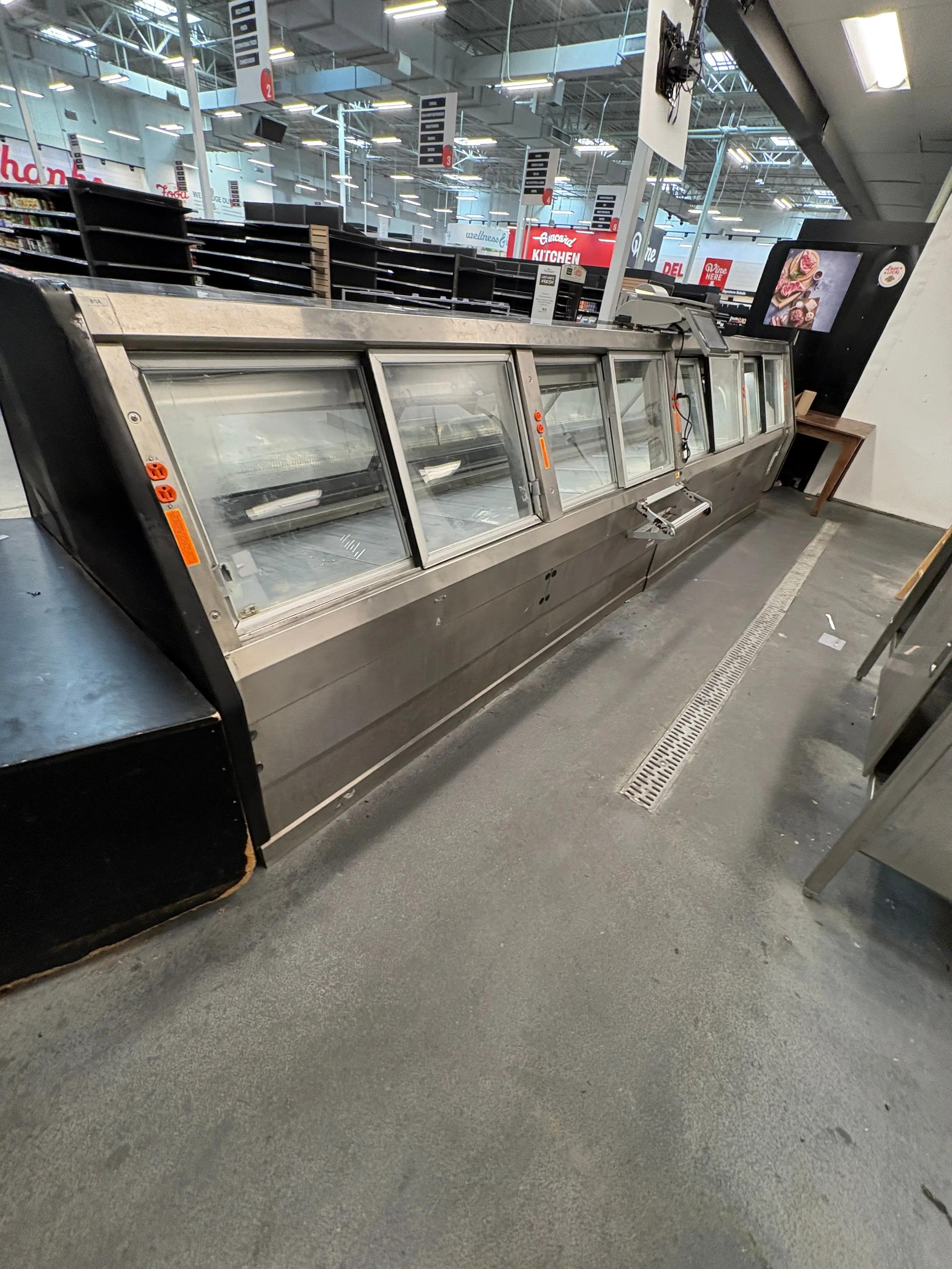 A commercial kitchen with stainless steel refrigeration units and shelving, part of a larger store environment with visible signage and overhead lighting fixtures.