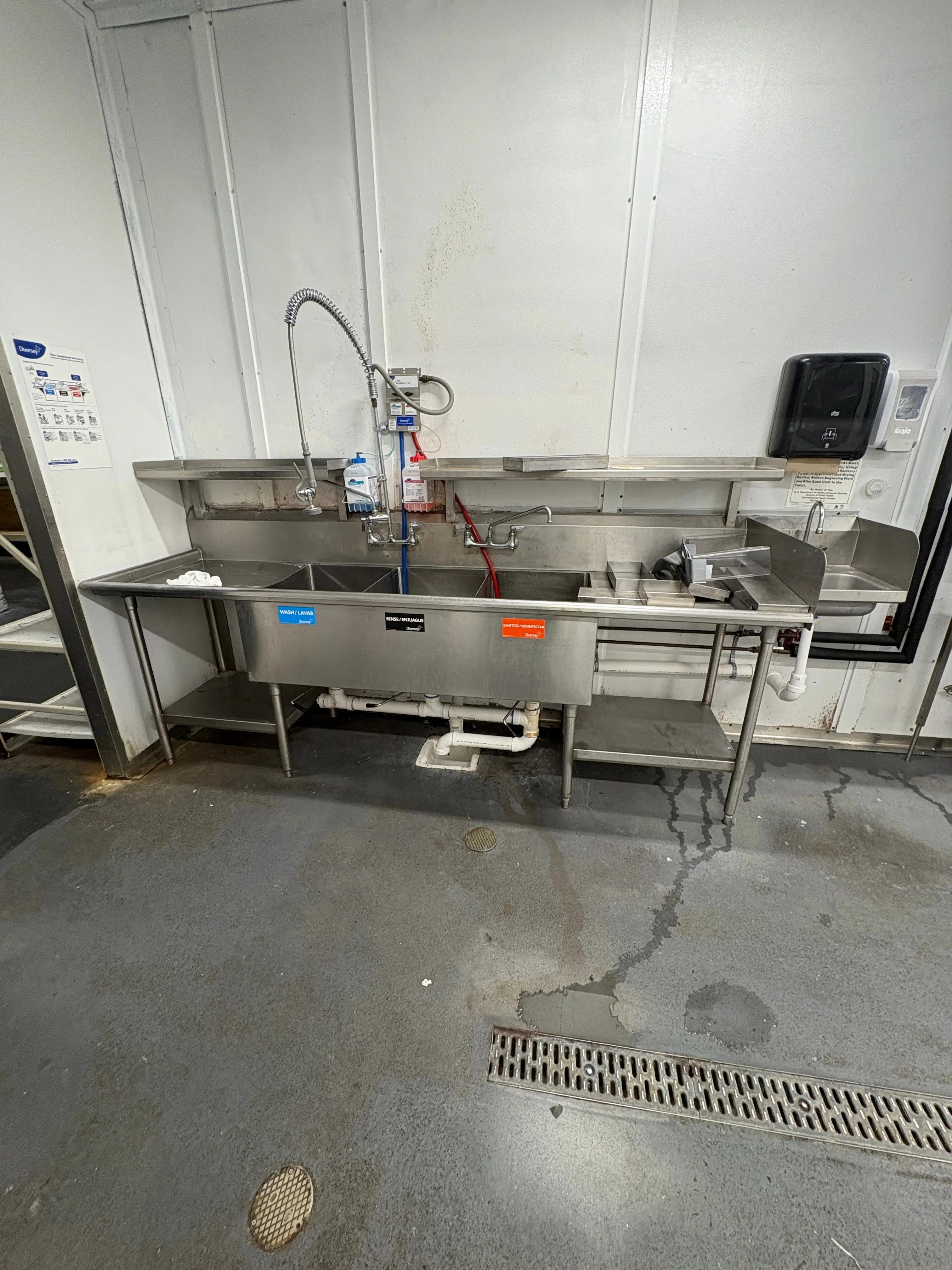 A commercial kitchen sink area with stainless steel fixtures, including a large sink, faucet, and soap dispensers. From an upcoming liquidation by Mid-America Store Fixtures