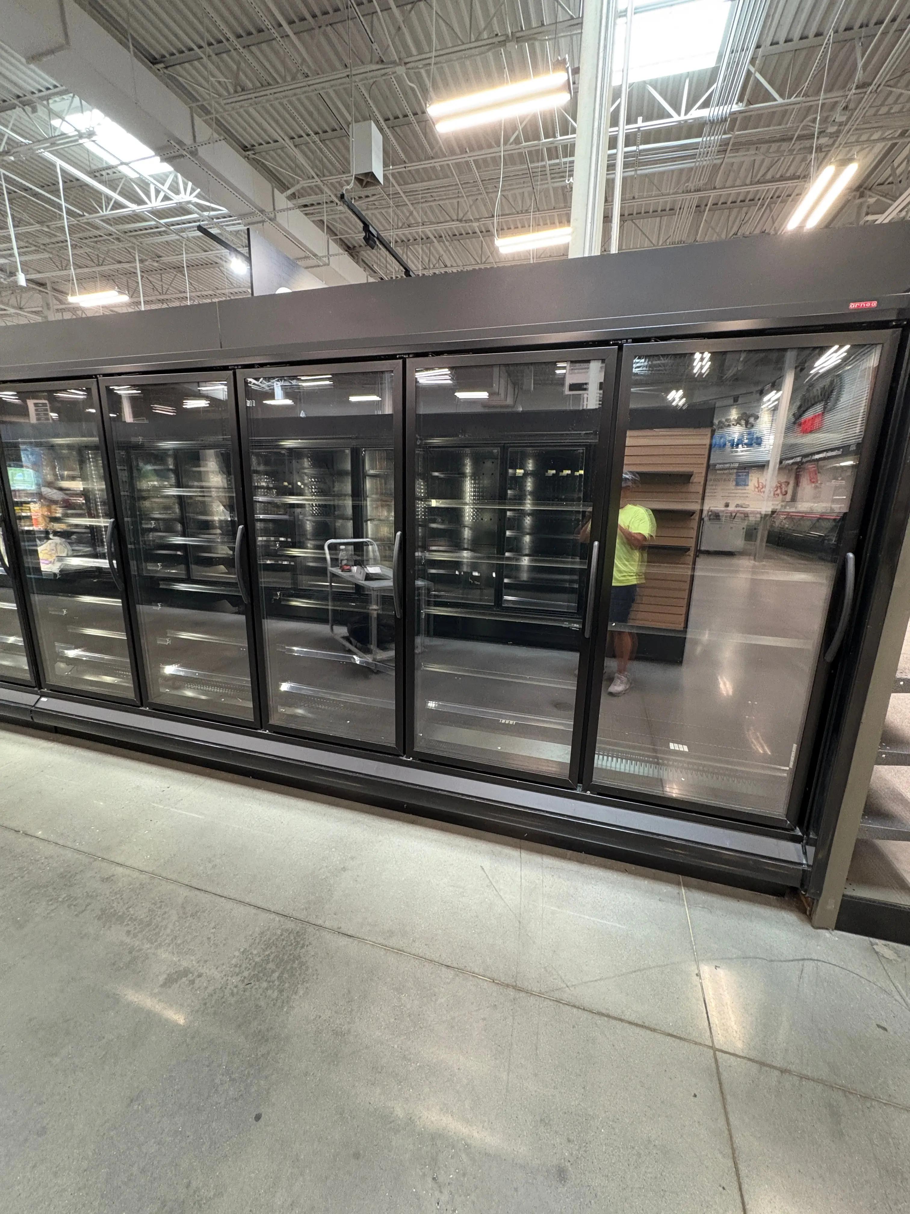 A row of commercial glass-door refrigerators in a store with bright overhead lighting. For sale from an upcoming liquidation with Mid-America Store Fixtures