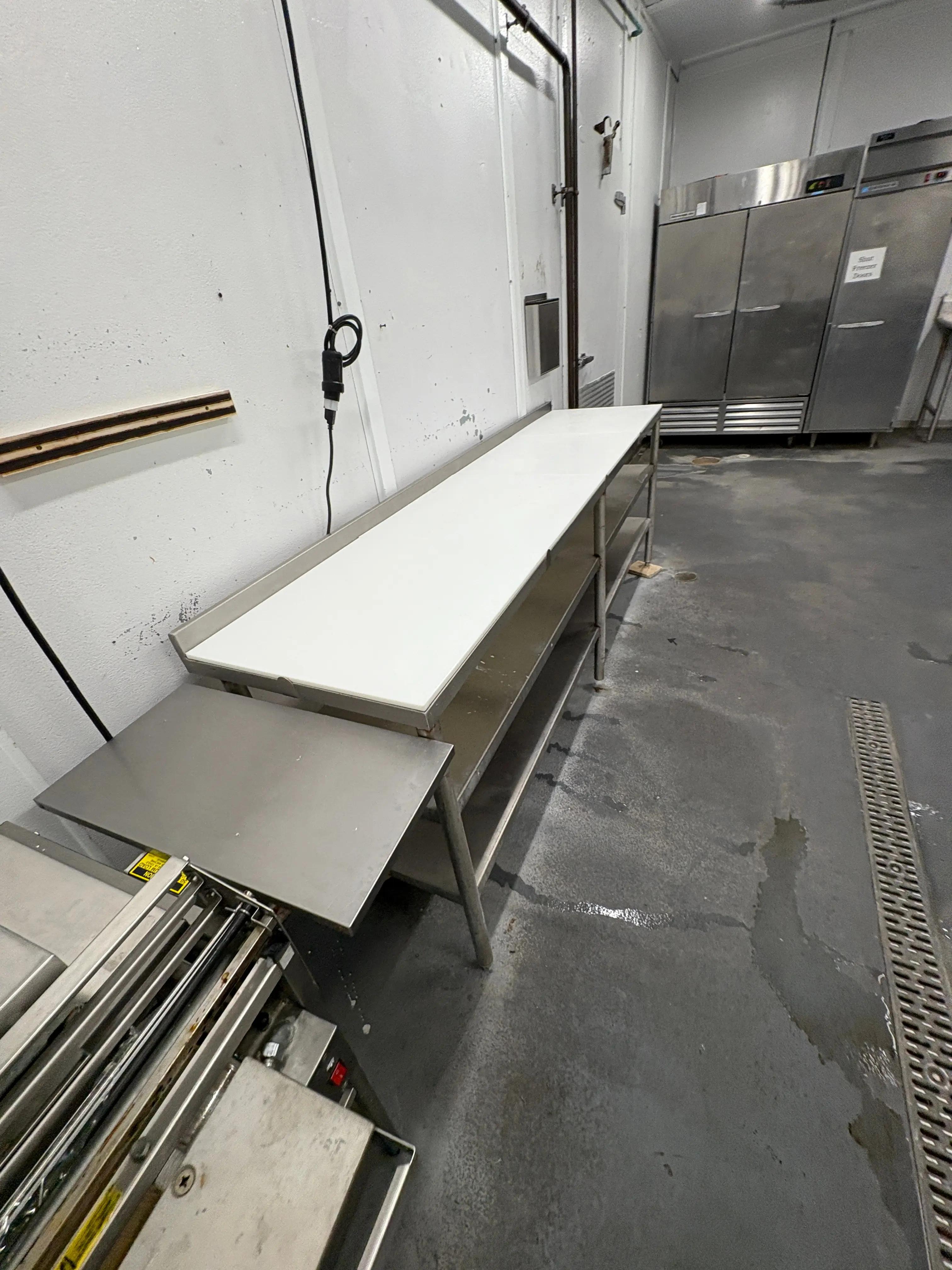 A commercial kitchen with a stainless steel table and an industrial refrigerator in the background, surrounded by a clean, gray concrete floor.