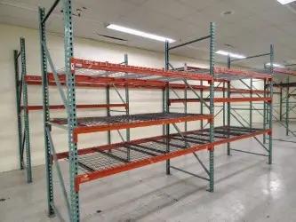 Empty industrial metal storage racks with red and green framework, installed in a warehouse space with a tiled floor and overhead lighting.