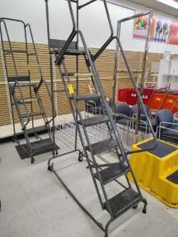 Industrial metal ladders with steps and handrails are displayed in a store setting, surrounded by chairs and shopping carts.