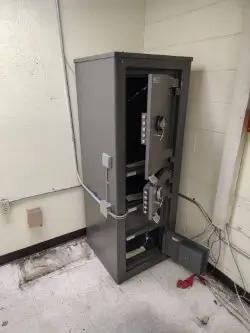 Open metal safe in a room corner with visible wiring and tiled floor, showcasing a security installation by Mid America.