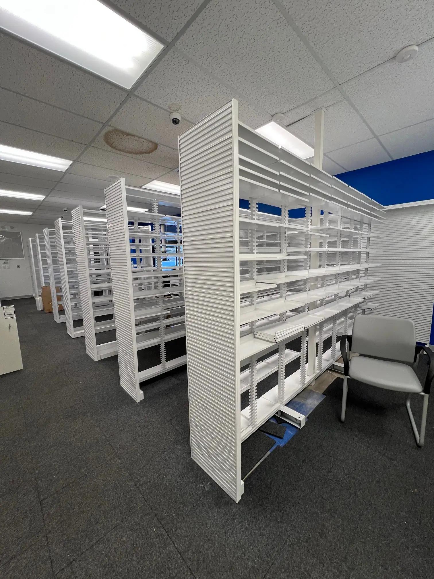 An office space with multiple white shelving units aligned in rows, gray carpet tiles on the floor, and ceiling lights. A single gray chair is partially visible.