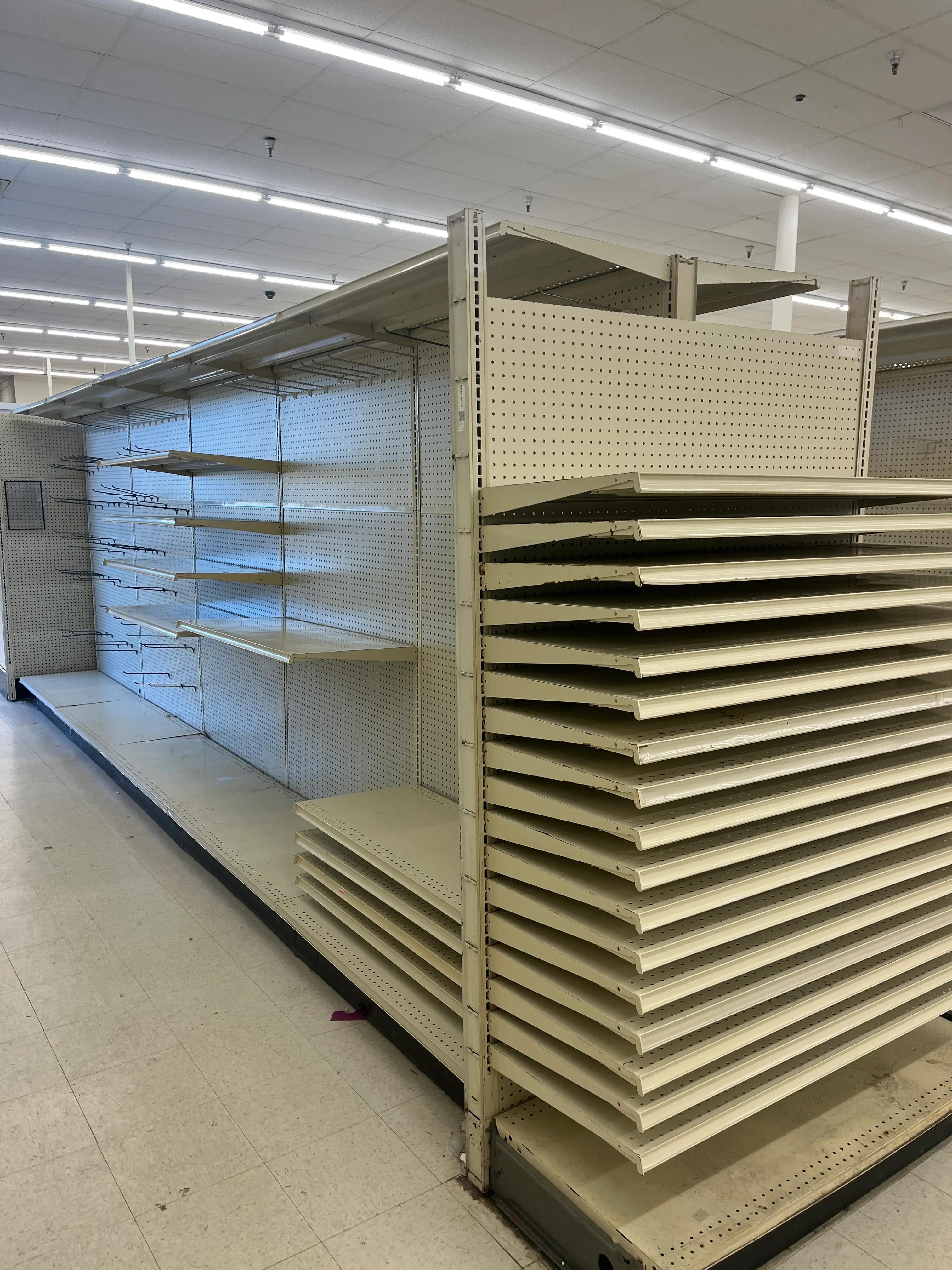 Empty retail store shelves with metal pegboard backing and stacked shelves on the side, illuminated by fluorescent lighting.