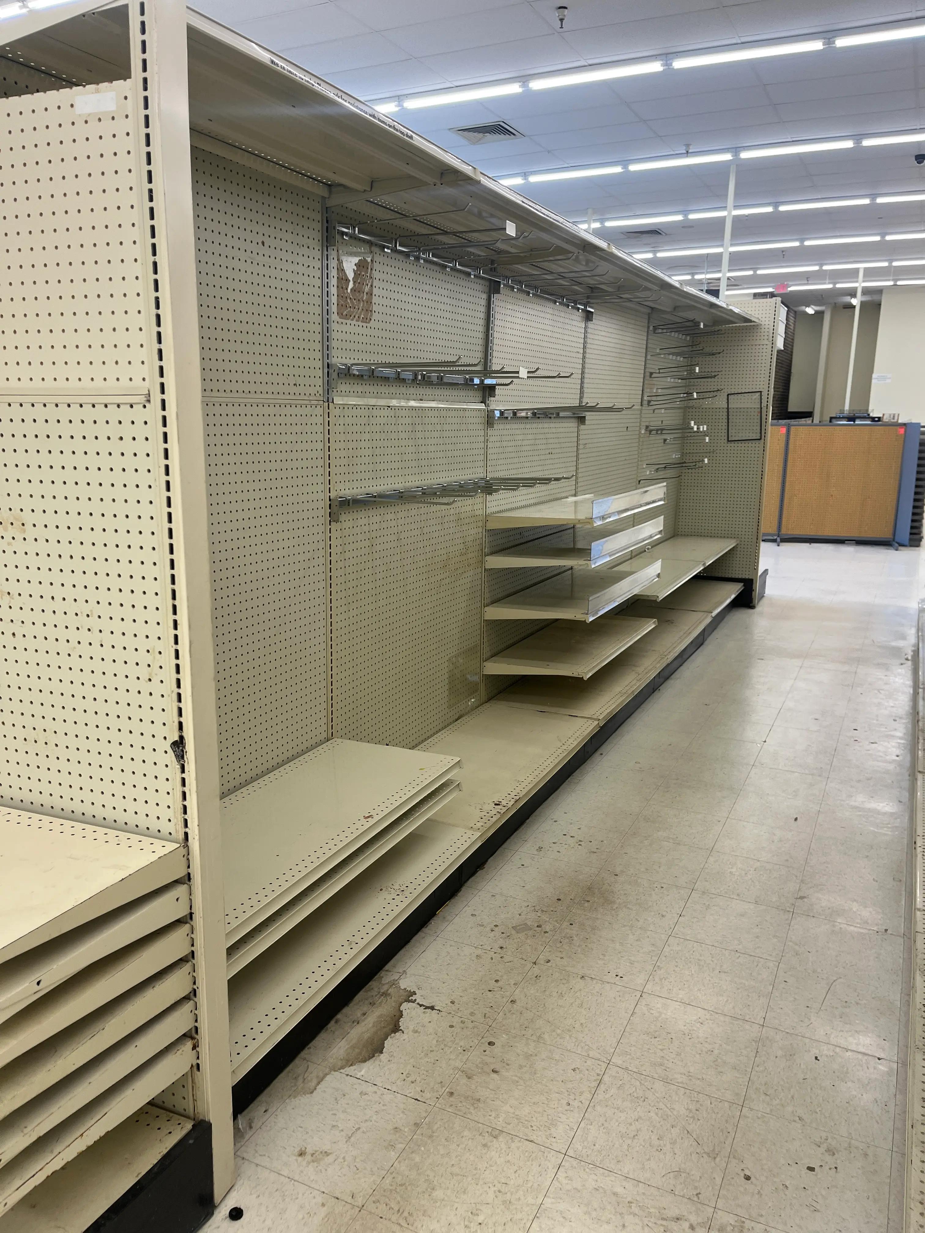 Empty retail shelving units with pegboard backing and metal brackets in a store. The layout is clean and organized, with a tiled floor and fluorescent lighting on the ceiling.