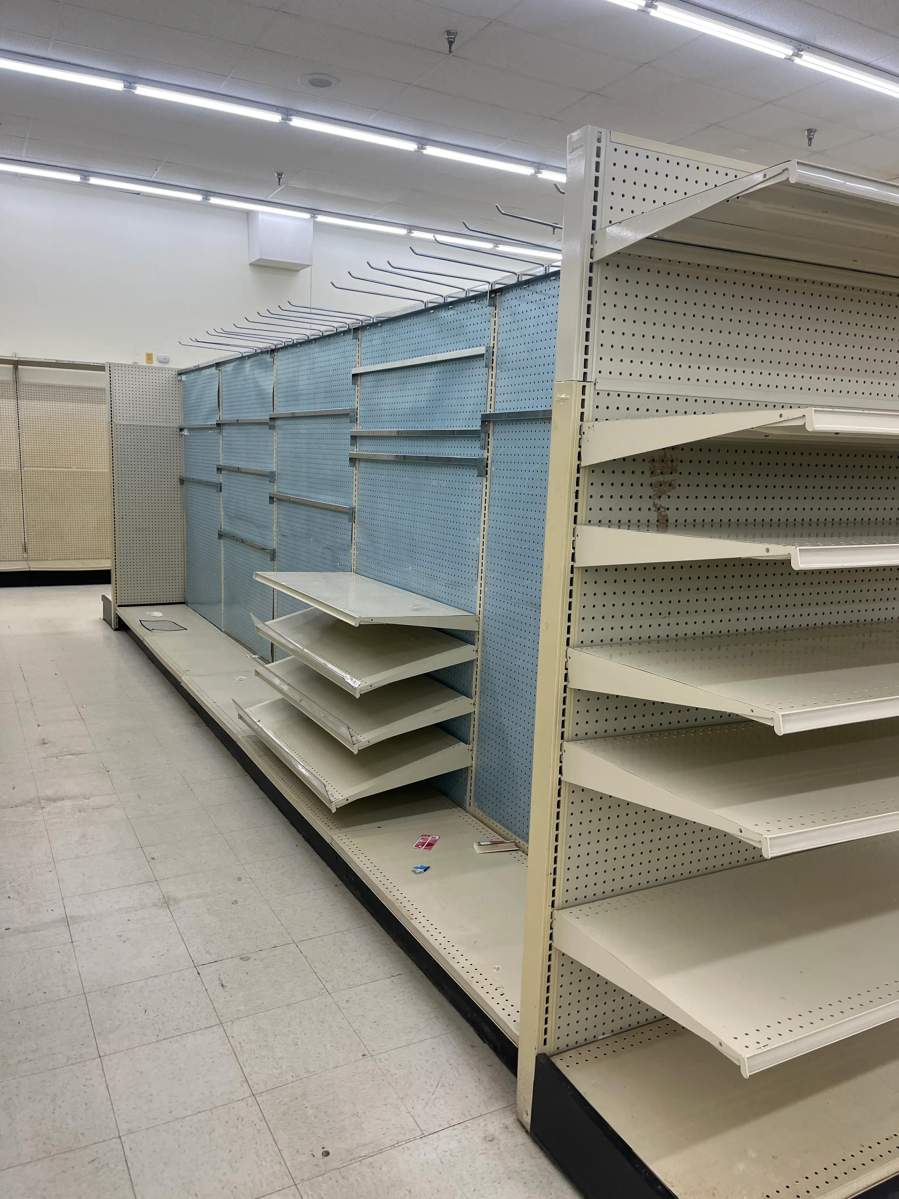Empty retail shelving units with pegboard backing and hooks in a store with fluorescent lighting and tiled flooring. Shelves are mostly bare, with a few scattered items on the bottom shelf.