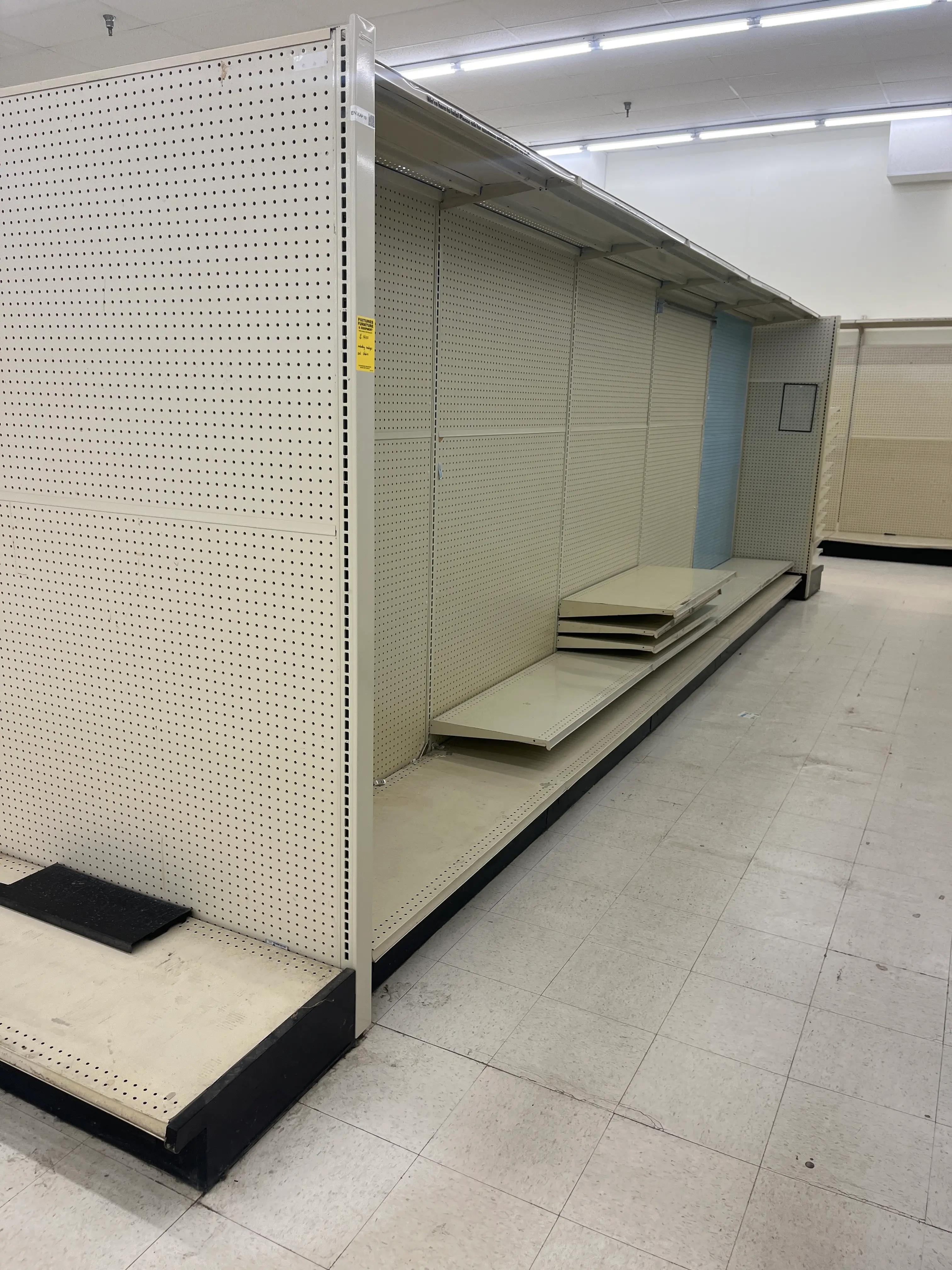 Empty retail shelving units with beige pegboard backing and disassembled shelves on the floor. The store features a tiled floor and fluorescent lighting overhead.