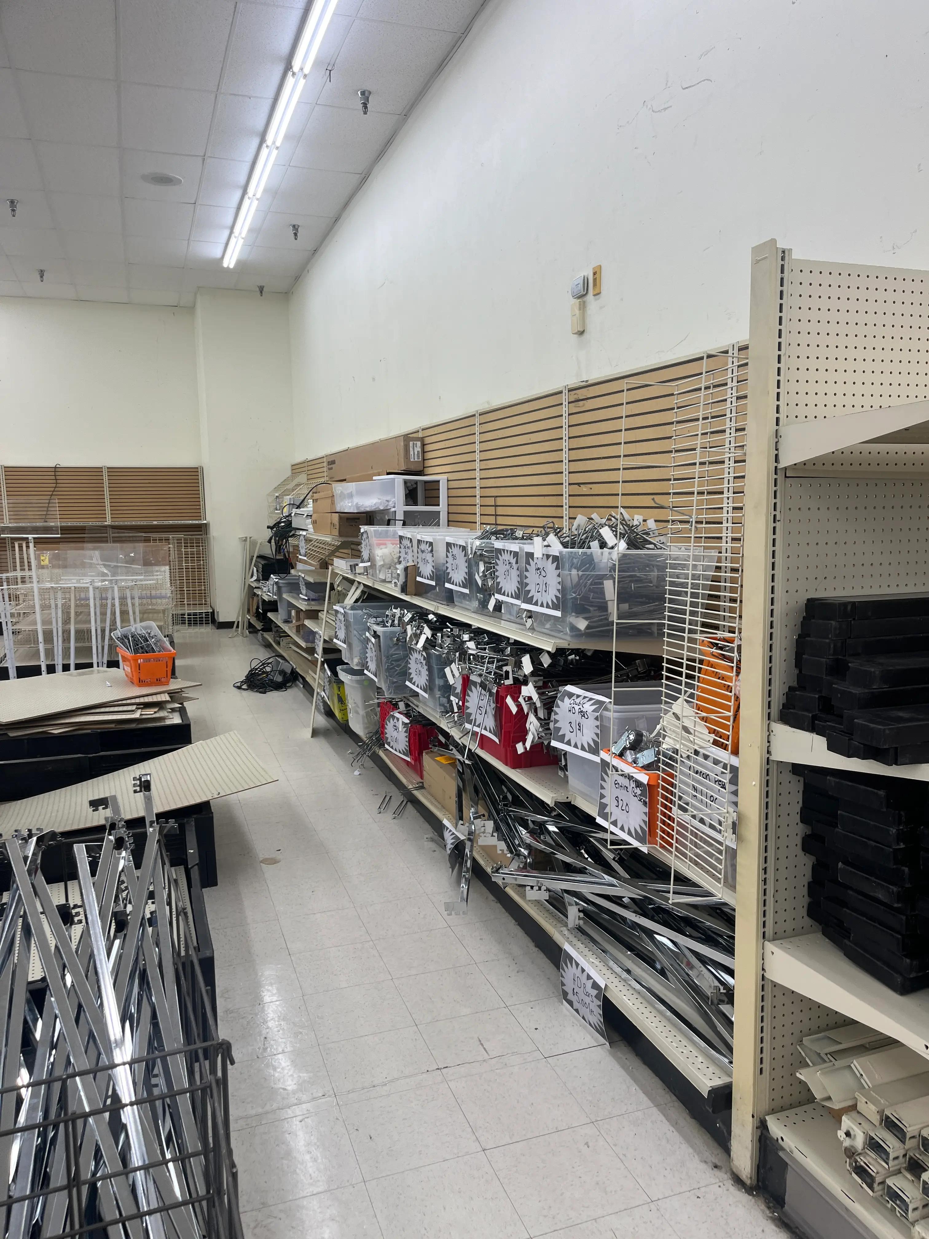 Store aisle with shelves holding metal and plastic parts, bins, and boxes. Tiled floor and fluorescent-lit ceiling. Shelves partially filled, suggesting restocking or work in progress.