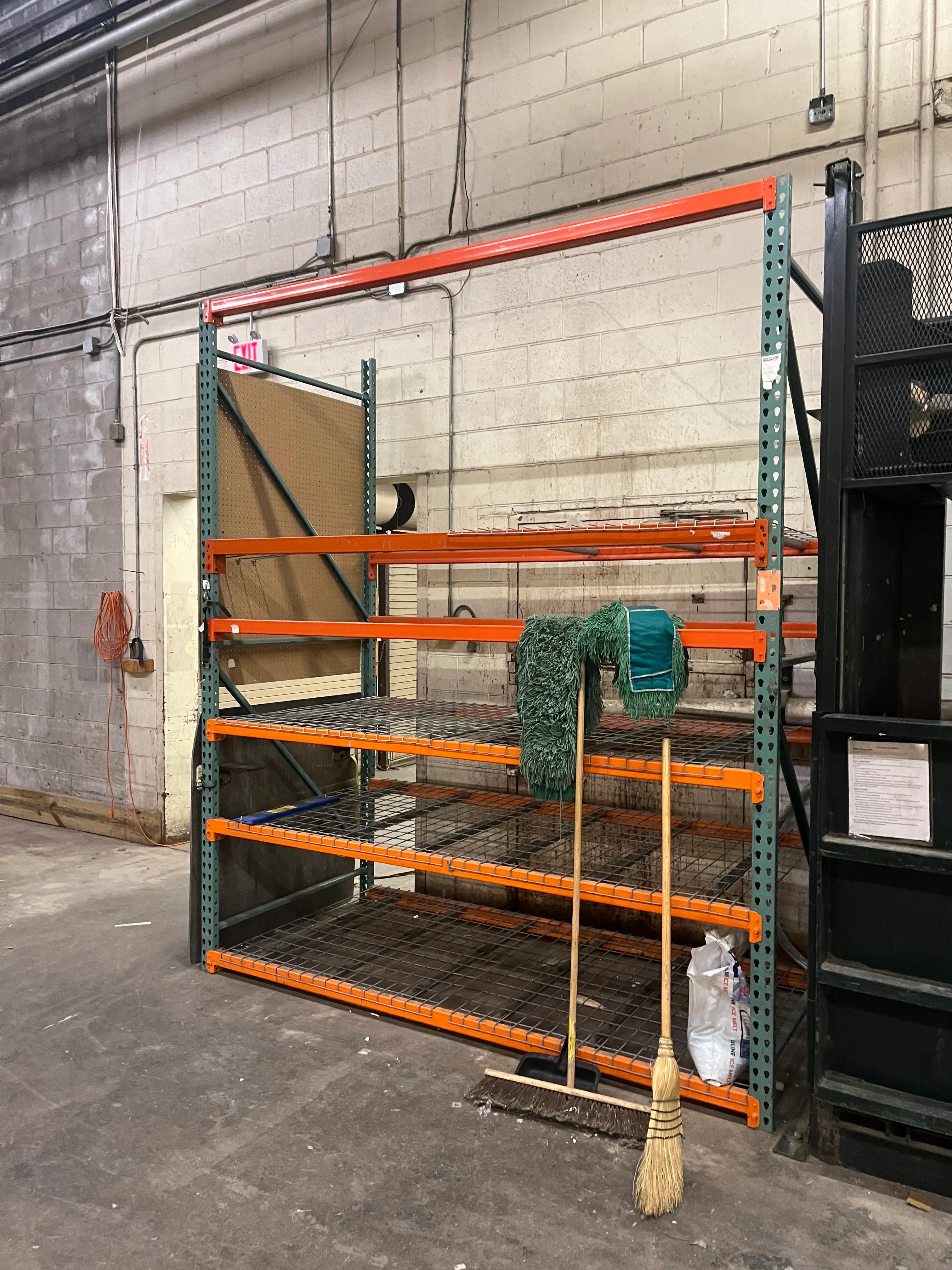 Industrial shelving unit with orange beams and green supports in a warehouse. Cleaning tools like a mop and broom are leaning against the shelves. An exit sign is visible on the wall behind.