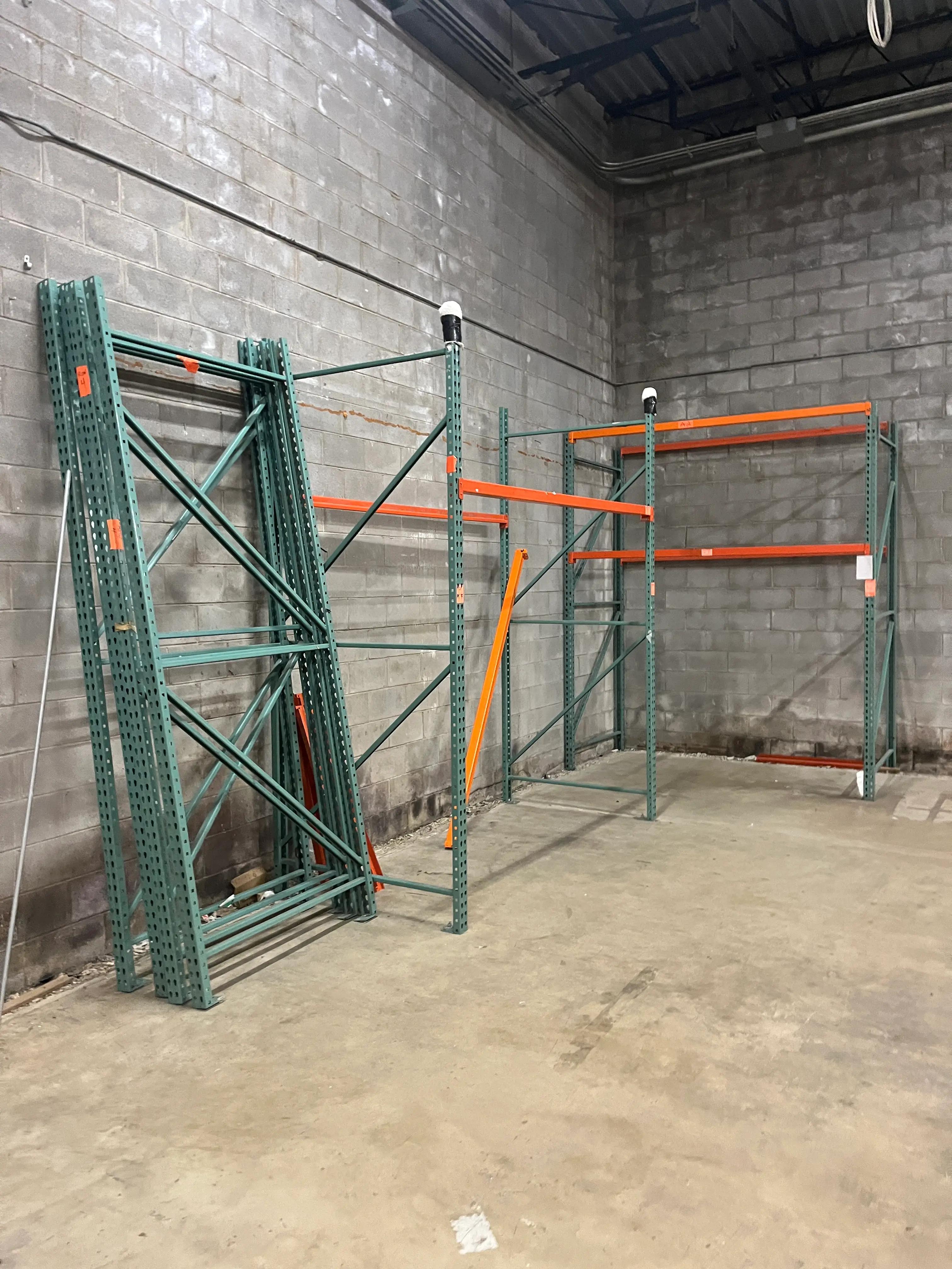 Industrial warehouse interior featuring partially assembled green and orange metal shelving units placed against a concrete block wall.