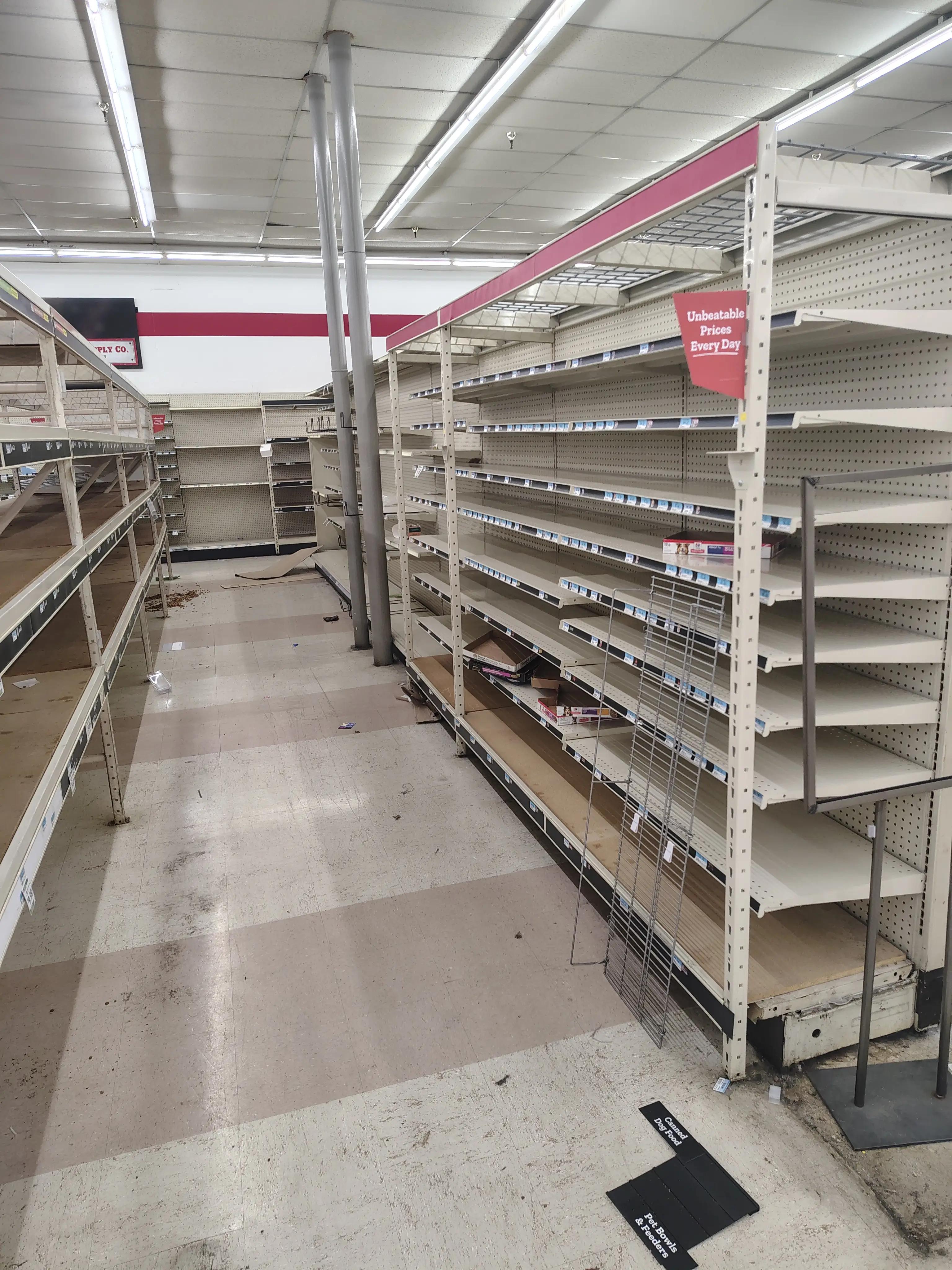 Empty shelves in a retail store with overhead fluorescent lighting and scattered items on the floor.