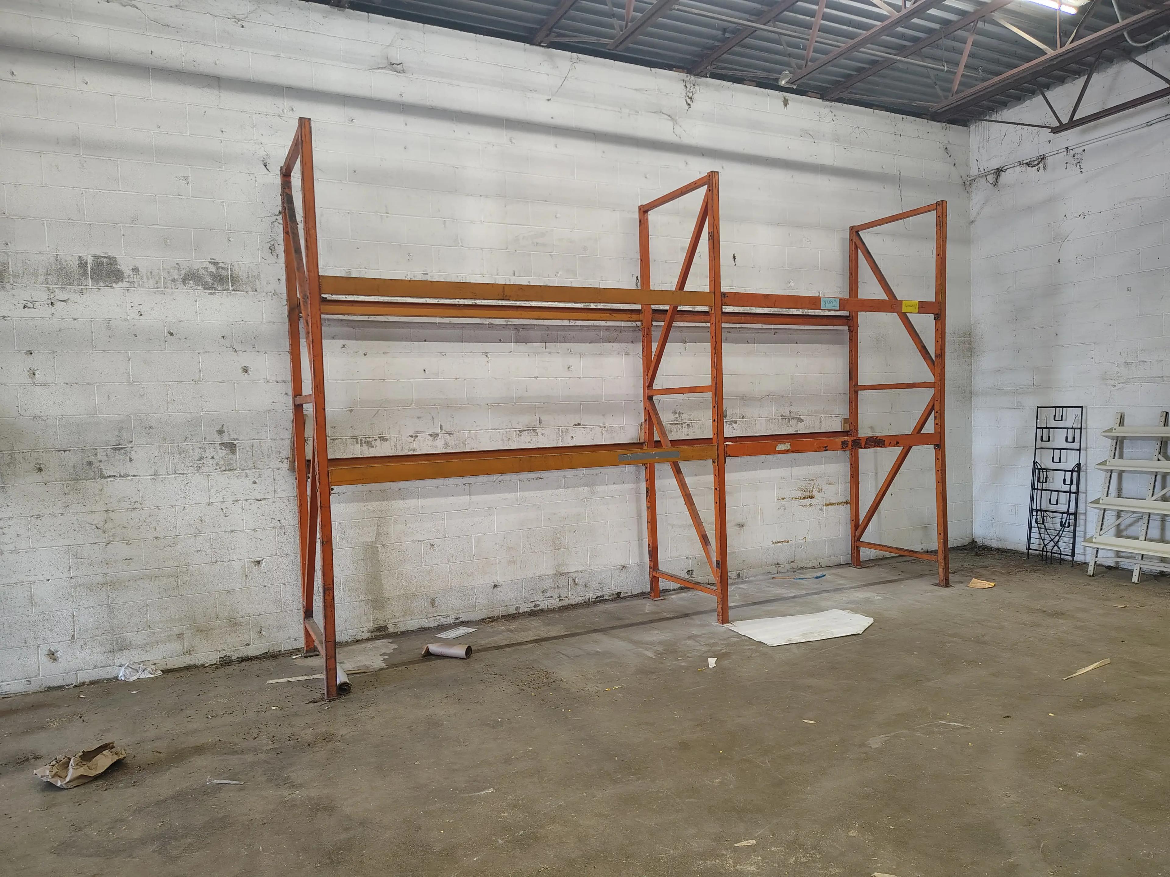 Empty industrial shelving unit against a white brick wall in a warehouse, with debris scattered on the concrete floor.