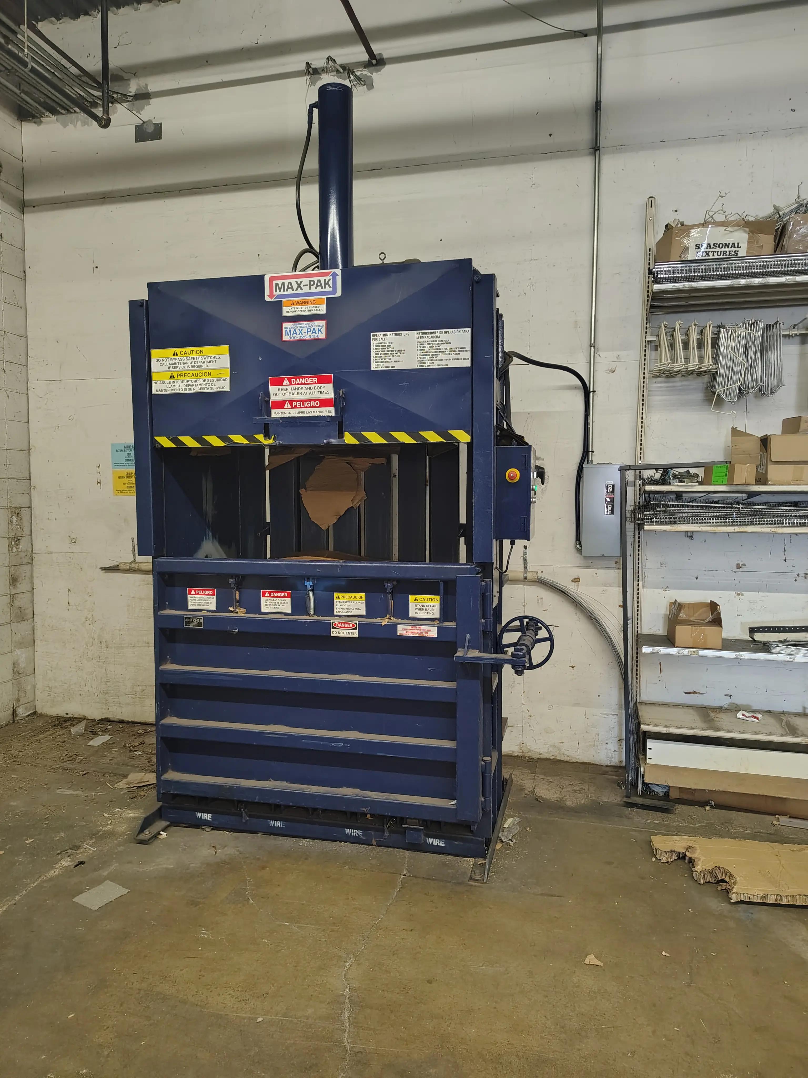 Industrial baler machine labeled "MAX-PAK" with safety warnings in English and Spanish, located in a warehouse. Shelves and cardboard are visible nearby.