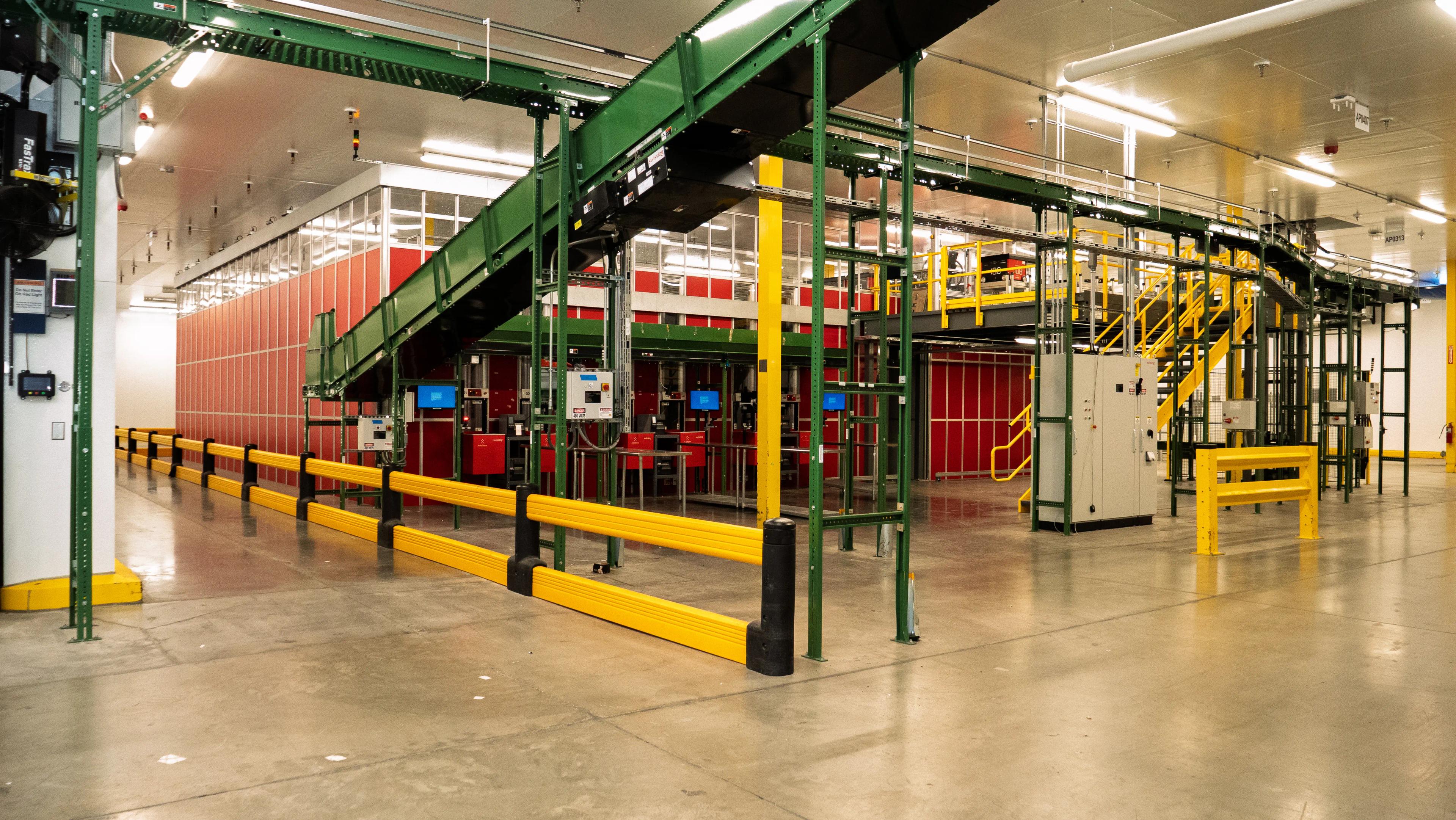 Interior view of an industrial facility featuring green conveyor belts, yellow barriers, and red storage units within a spacious warehouse setting.