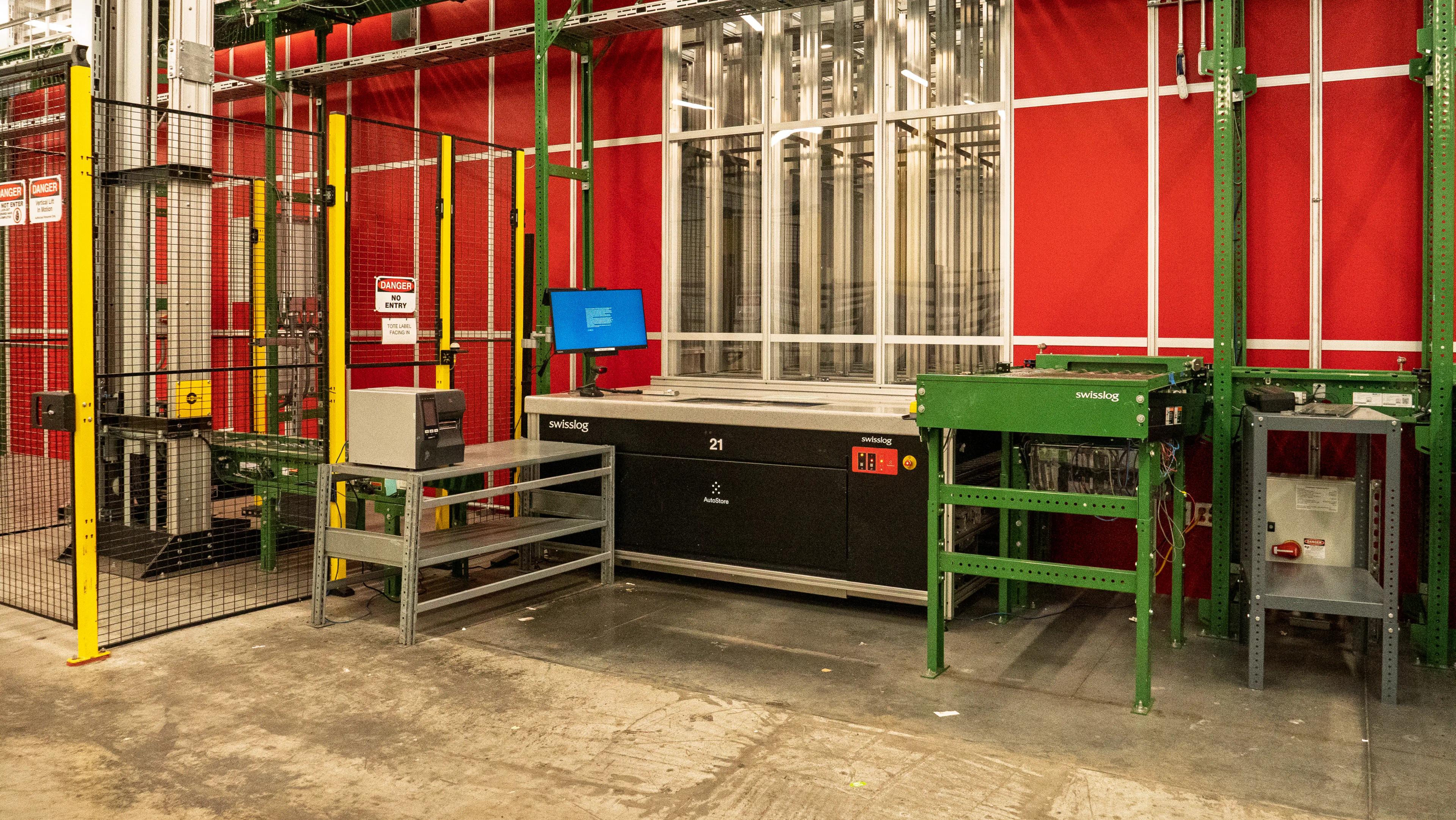 Industrial setup featuring automated machinery with red and green structural components, a safety fence, and a computer display.