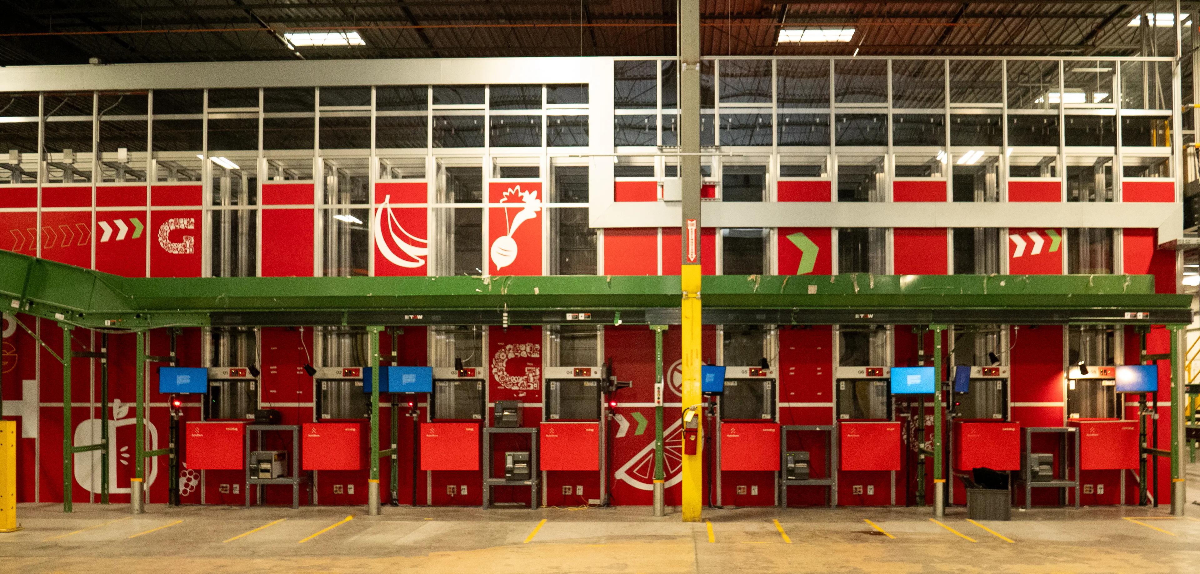 Interior of a warehouse featuring red modular storage units with a green conveyor system above. The storage units are adorned with white graphics of fruits and vegetables.