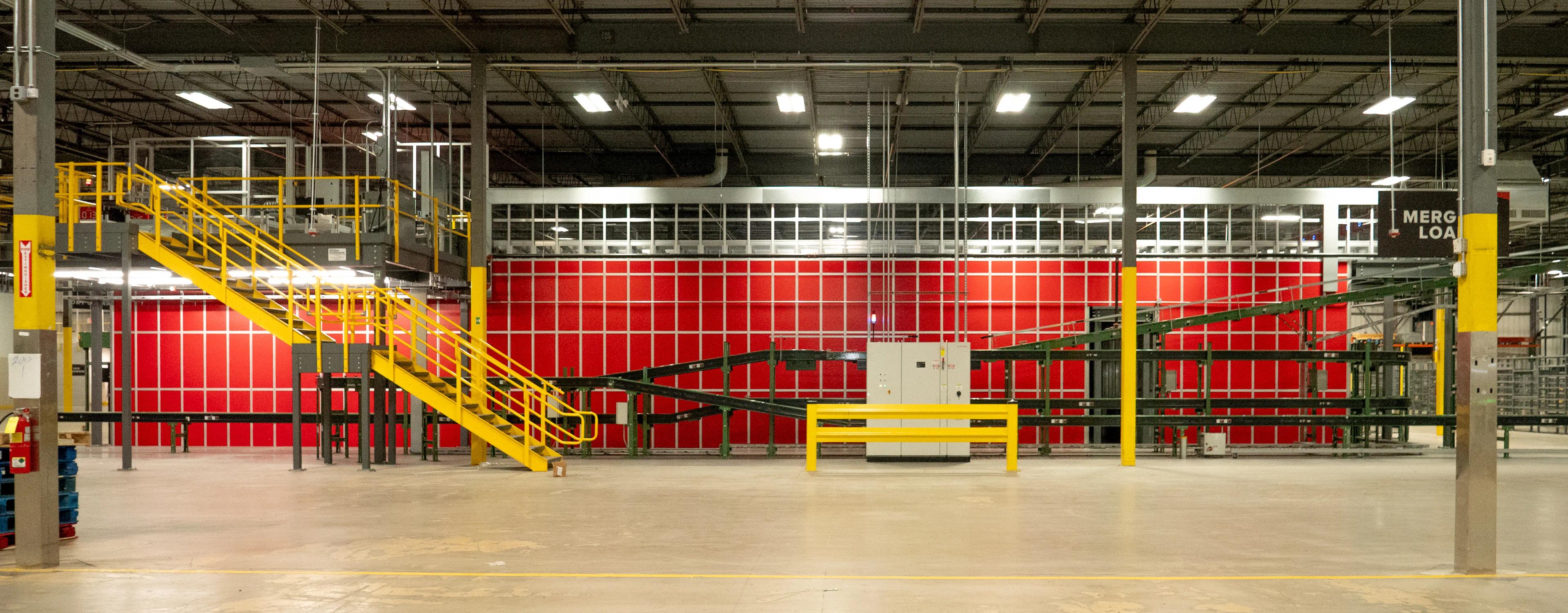 Interior of a large industrial warehouse with yellow railings, a conveyor system, and a bright red wall in the background.