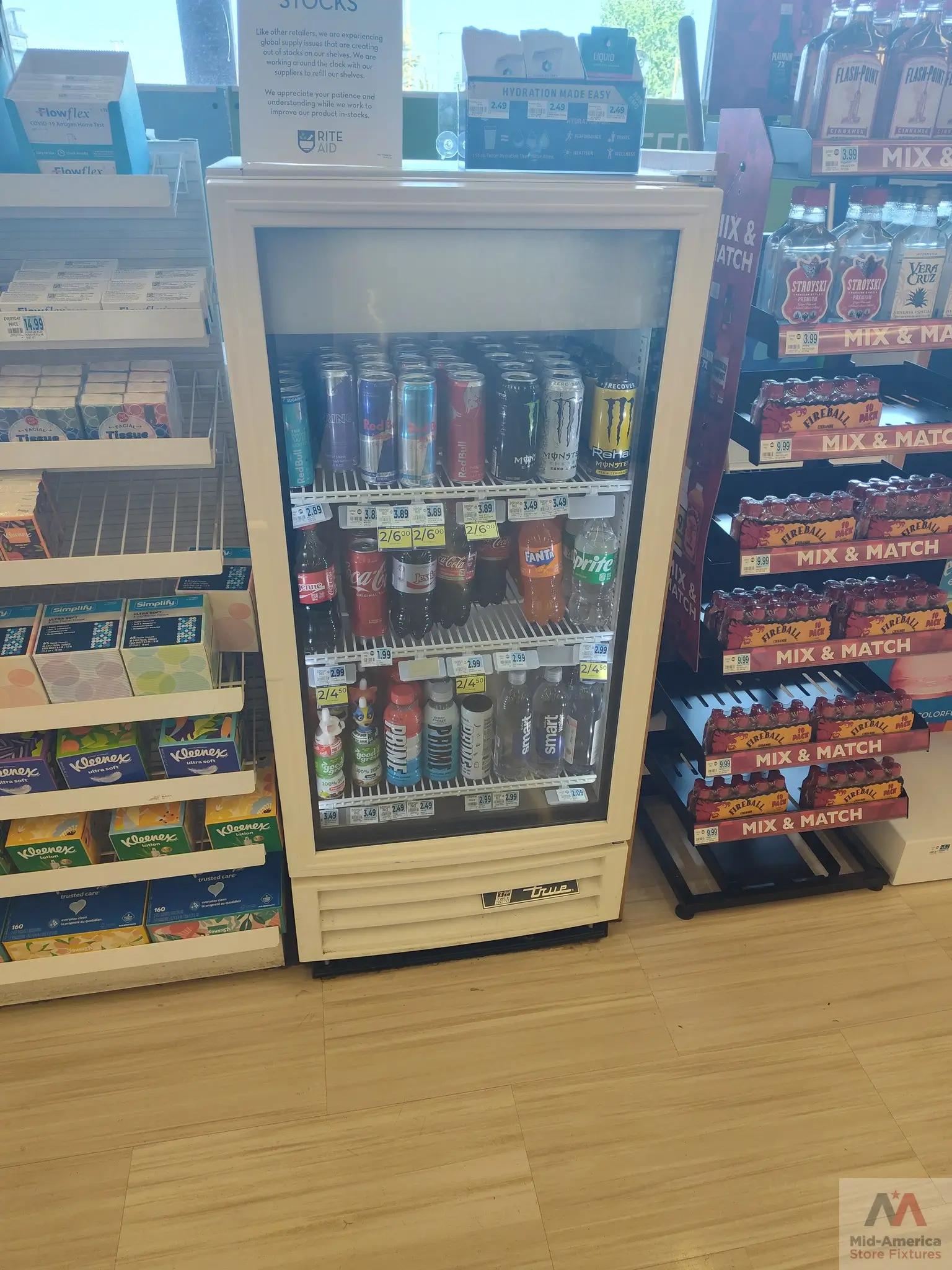 Store display with a refrigerated section containing energy drinks and sodas. Nearby shelves hold Kleenex tissues and Flowflex COVID-19 tests. A sign mentions global supply issues. Mid-America Store Fixtures logo is visible.