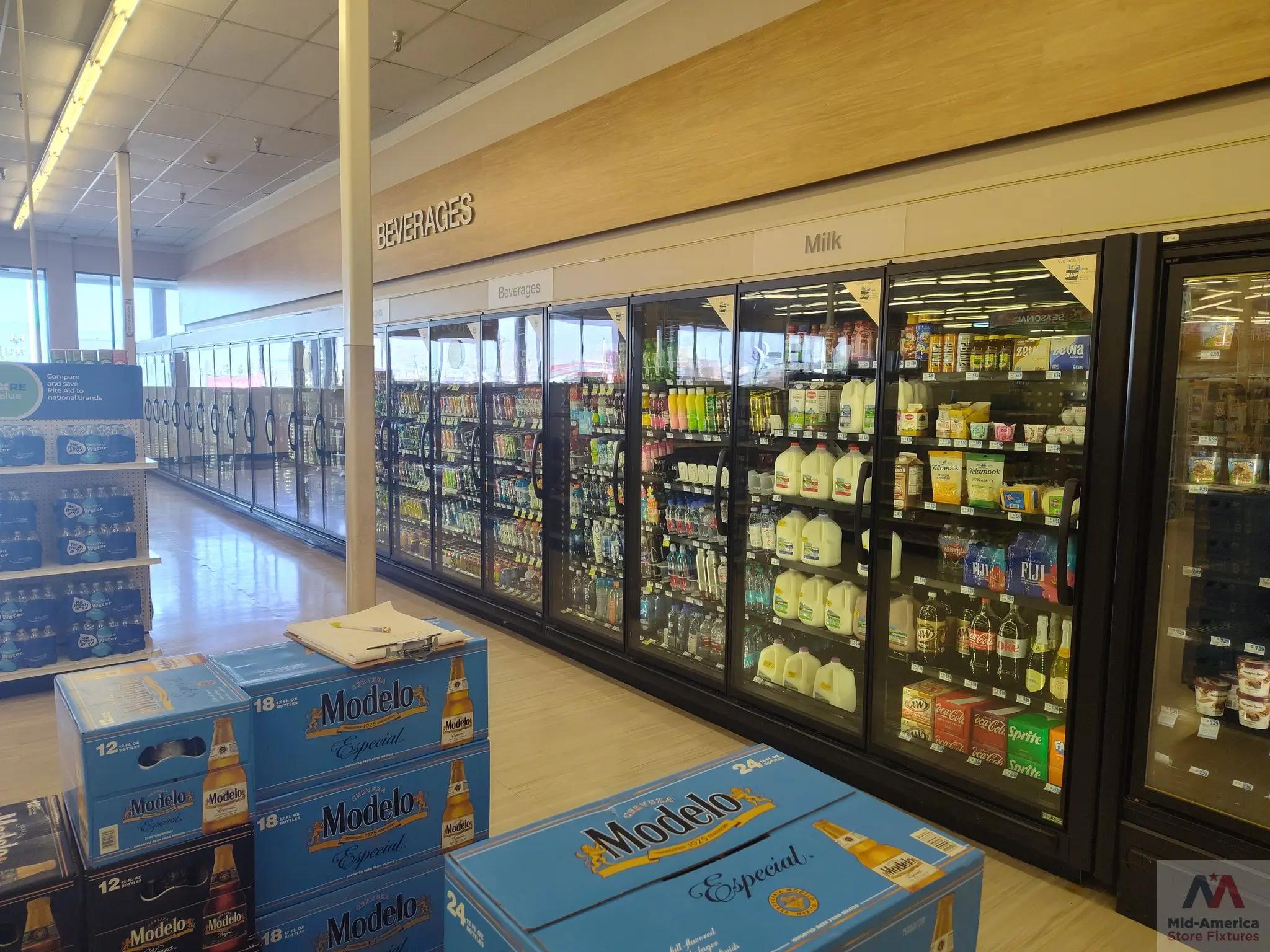 A grocery store aisle with refrigerated sections labeled "Beverages" and "Milk," featuring drinks like milk and soda. Boxes of Modelo Especial beer are stacked in the foreground. The Mid-America Store Fixtures logo is visible.