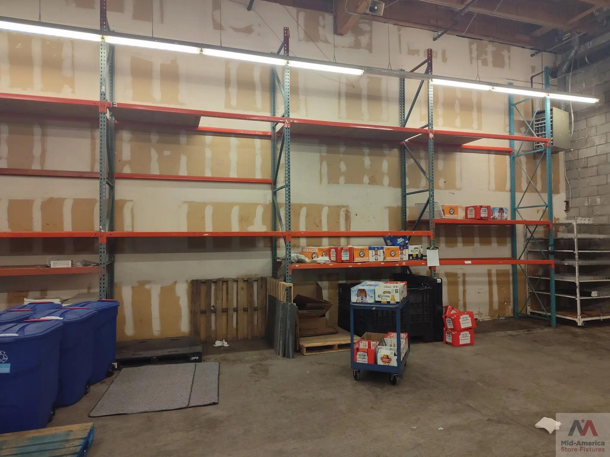 Interior of a warehouse with empty metal shelves, a few boxes of Corona beer, and blue recycling bins. The wall has patchy paint, and the floor is concrete. A logo for Mid-America Store Fixtures is visible in the corner.