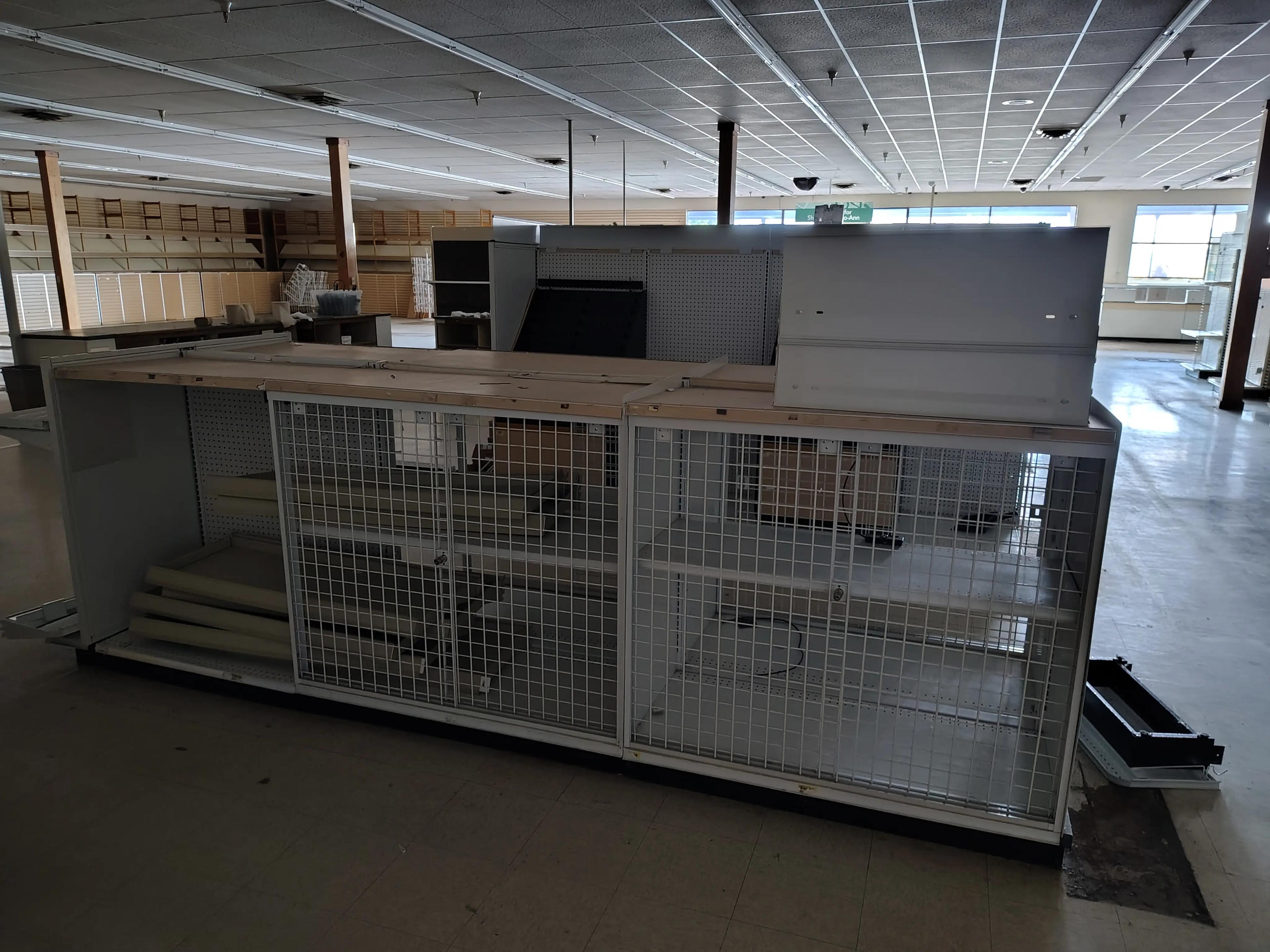 Empty retail space showing display shelving units and a grid display case, with overhead lighting and tiled floors.