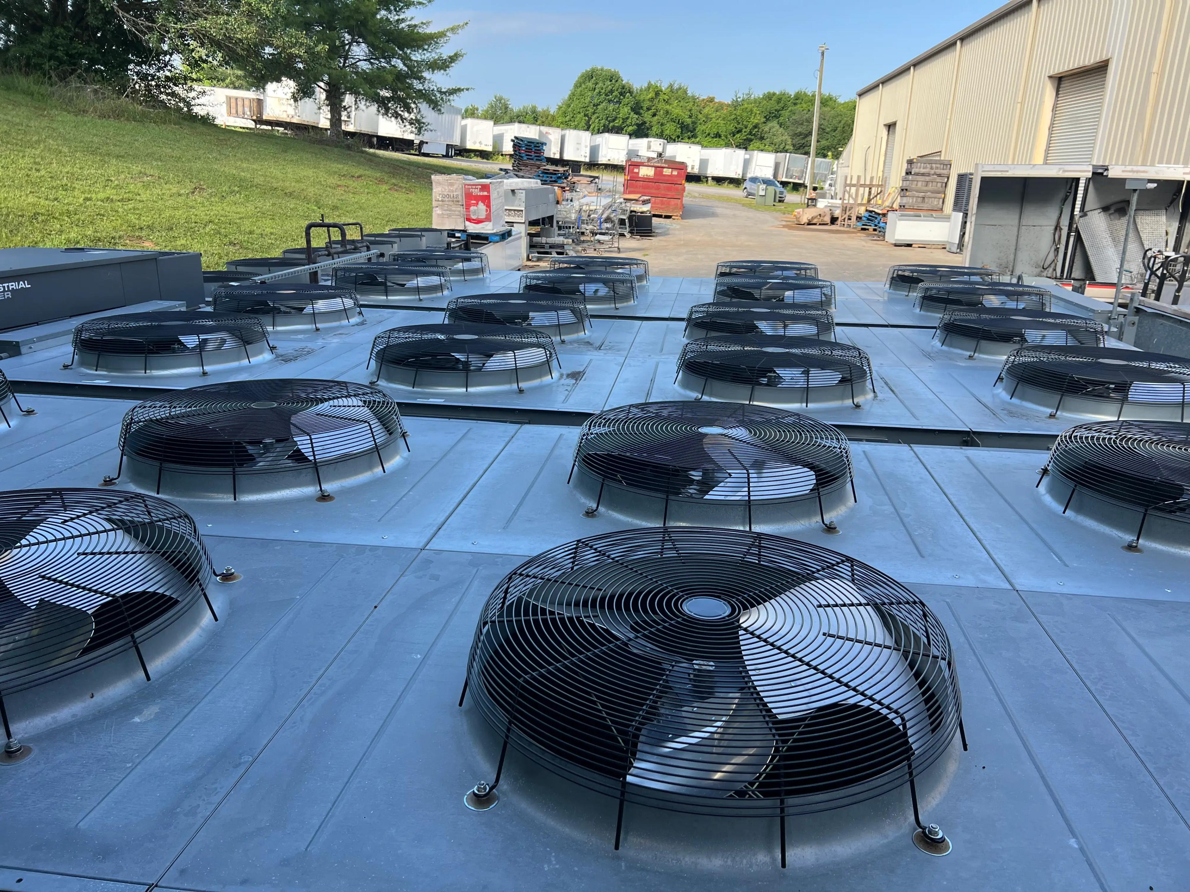 A series of industrial air conditioning units with fans on a metallic surface, located outdoors near a warehouse.