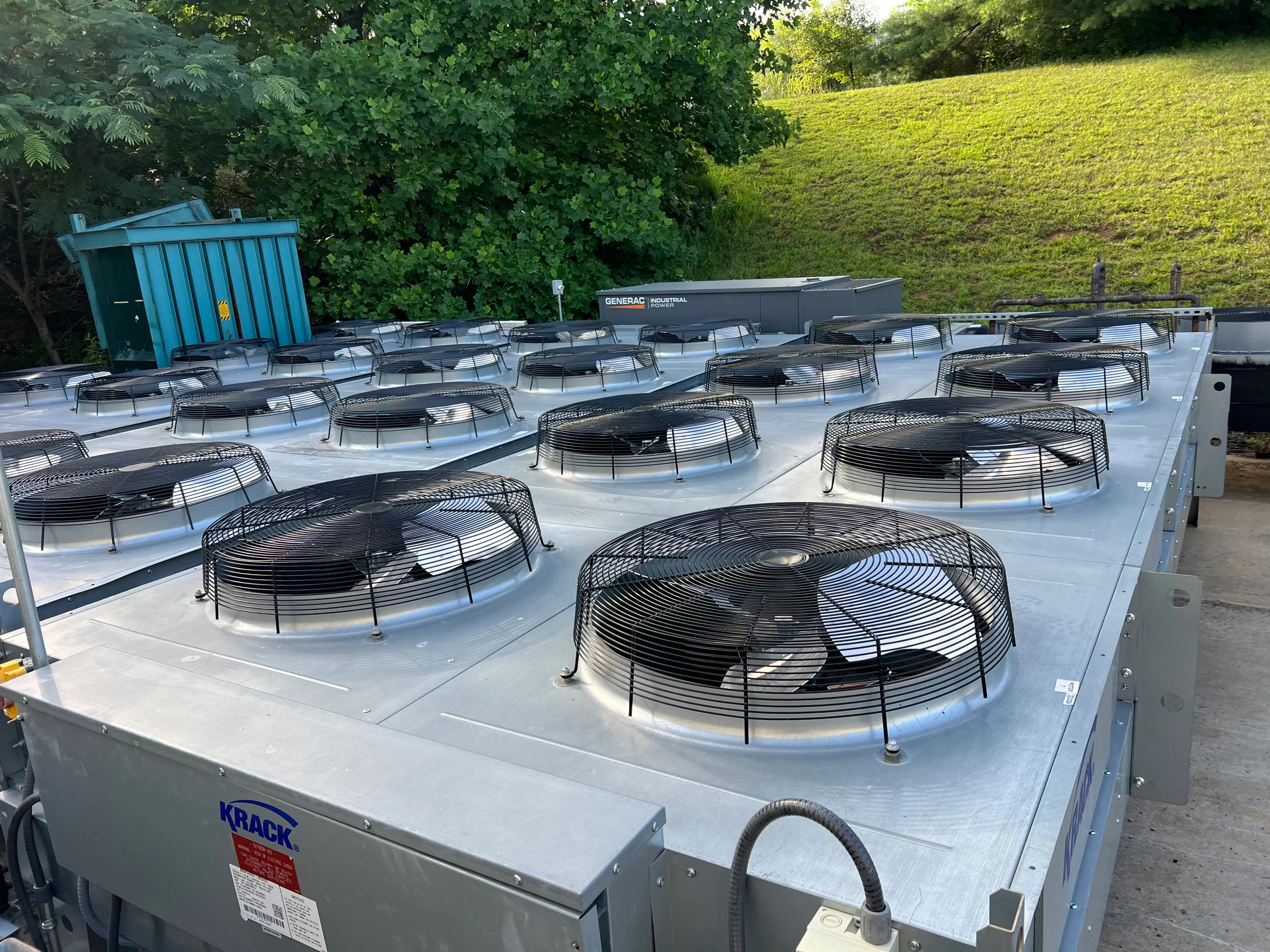 Industrial HVAC cooling units with multiple fans installed outdoors beside a hill and green trees.