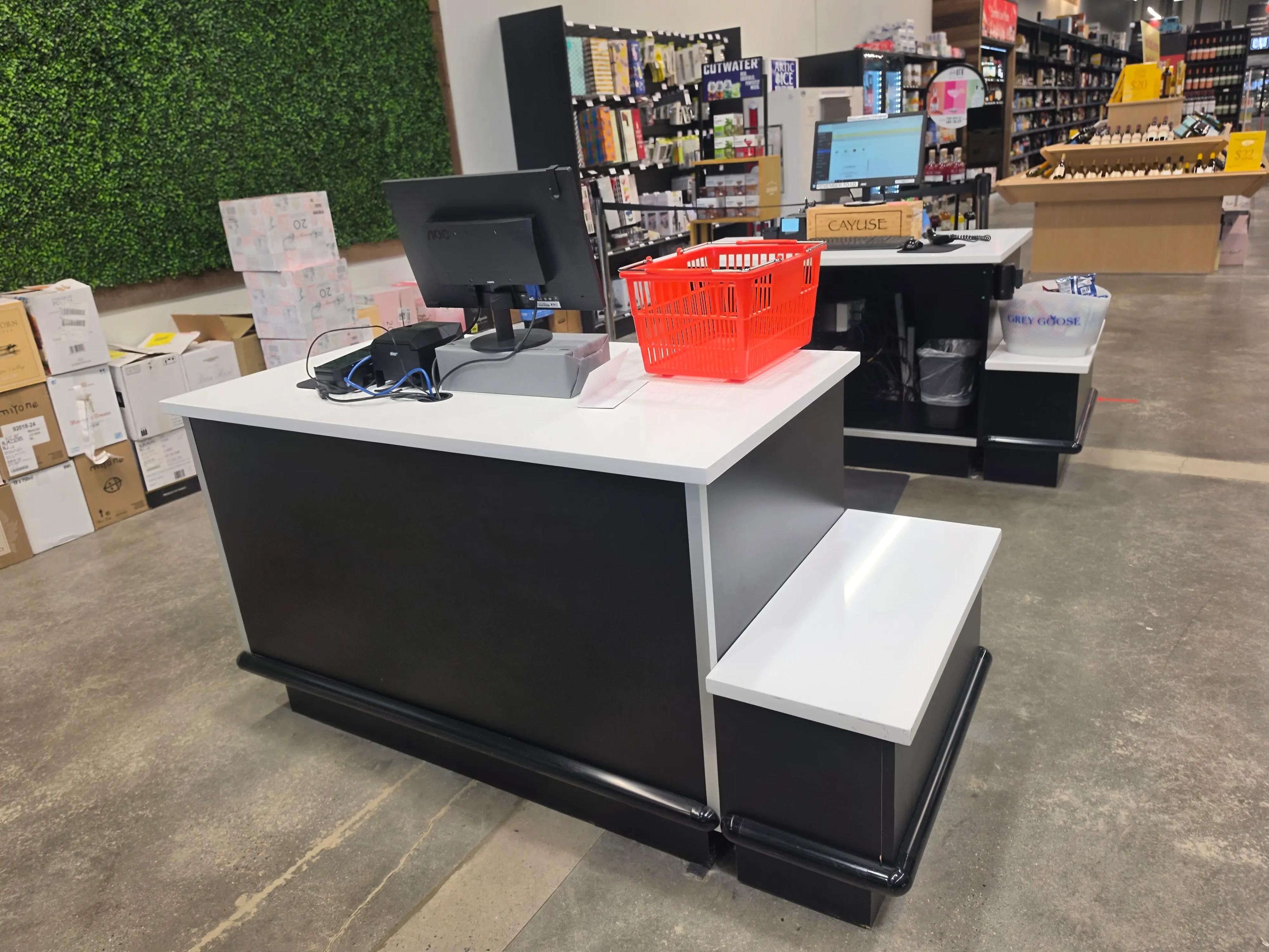 Checkout counter in a retail store featuring a computer, red basket, and various products on shelves. Boxes are stacked nearby against a green wall.