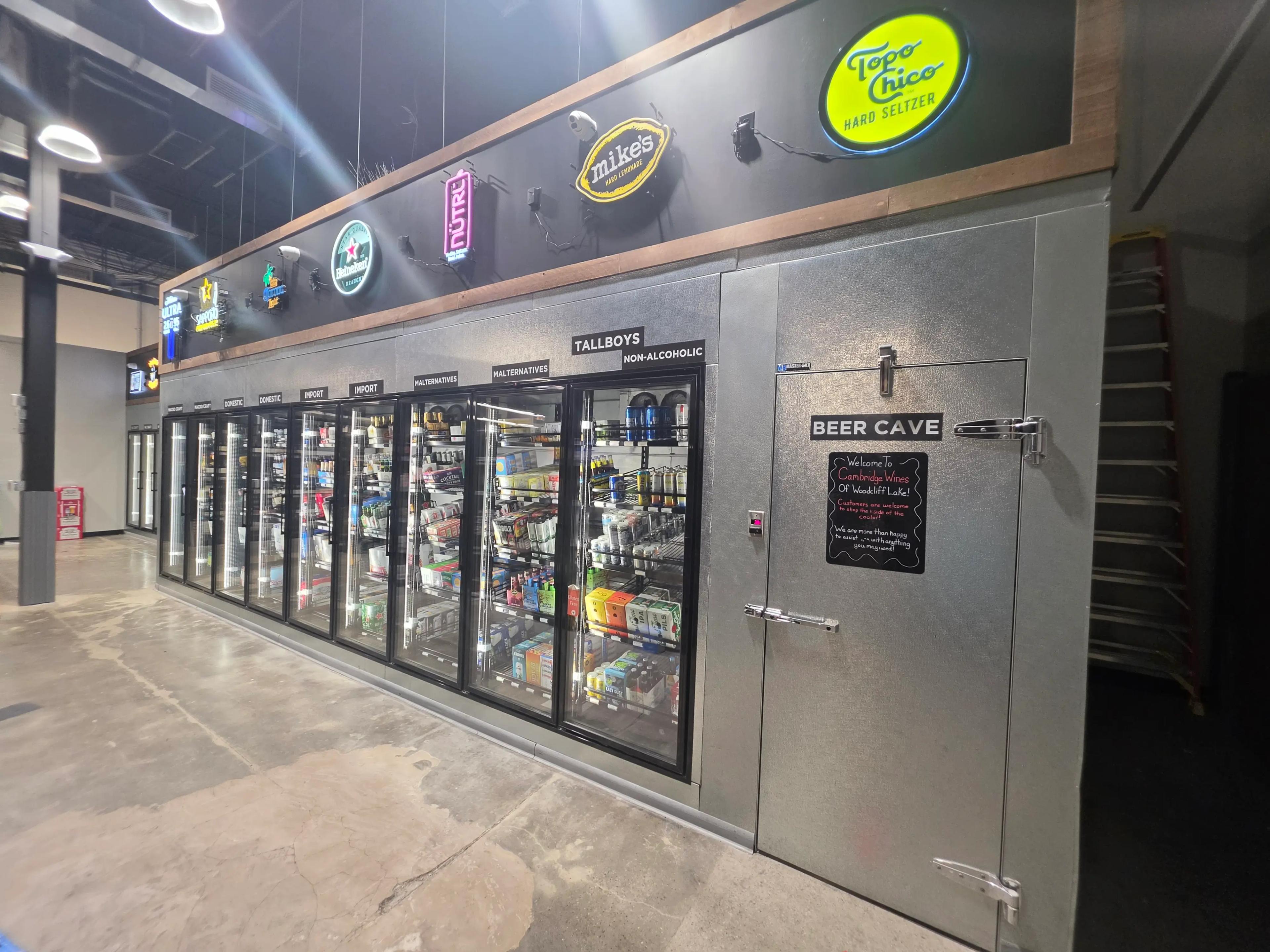 Refrigerated section in a store featuring beverage brands like Topo Chico, Mike's Hard Lemonade, and Heineken. A sign reads "Beer Cave" and "Welcome To Cambridge Wines Of Woodcliff Lake!" with a note about customer assistance.